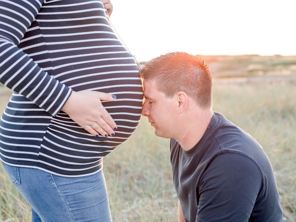 A tender maternity moment captured outdoors with a striped shirt and gentle belly touch in a natural setting.