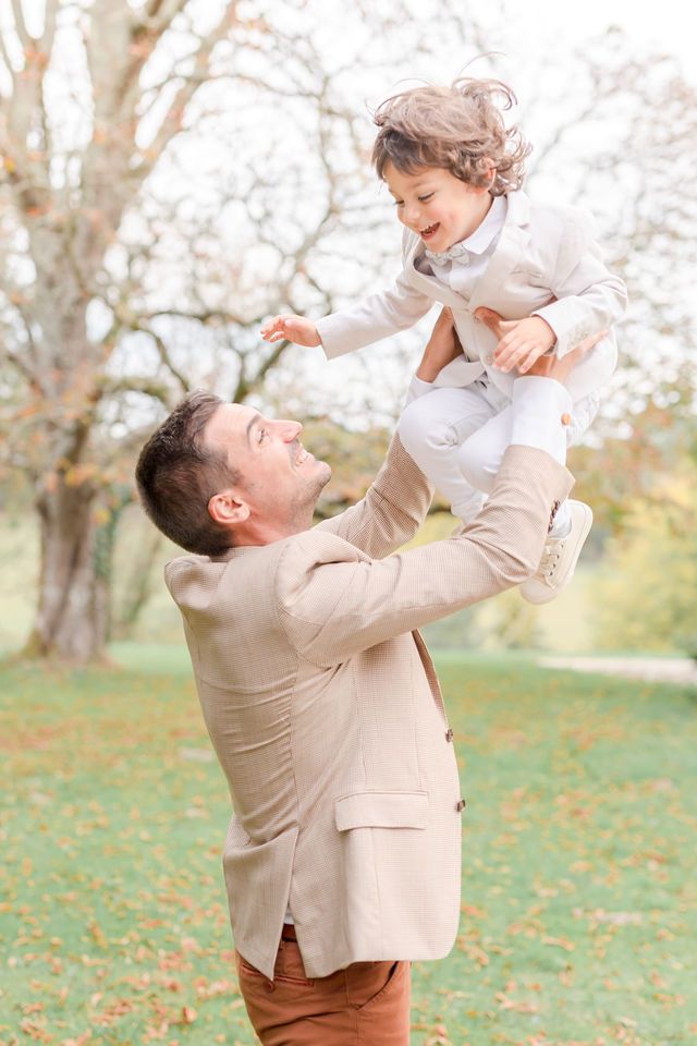 Playful outdoor moment with beige-suited figure lifting small child in white outfit among autumn trees.
