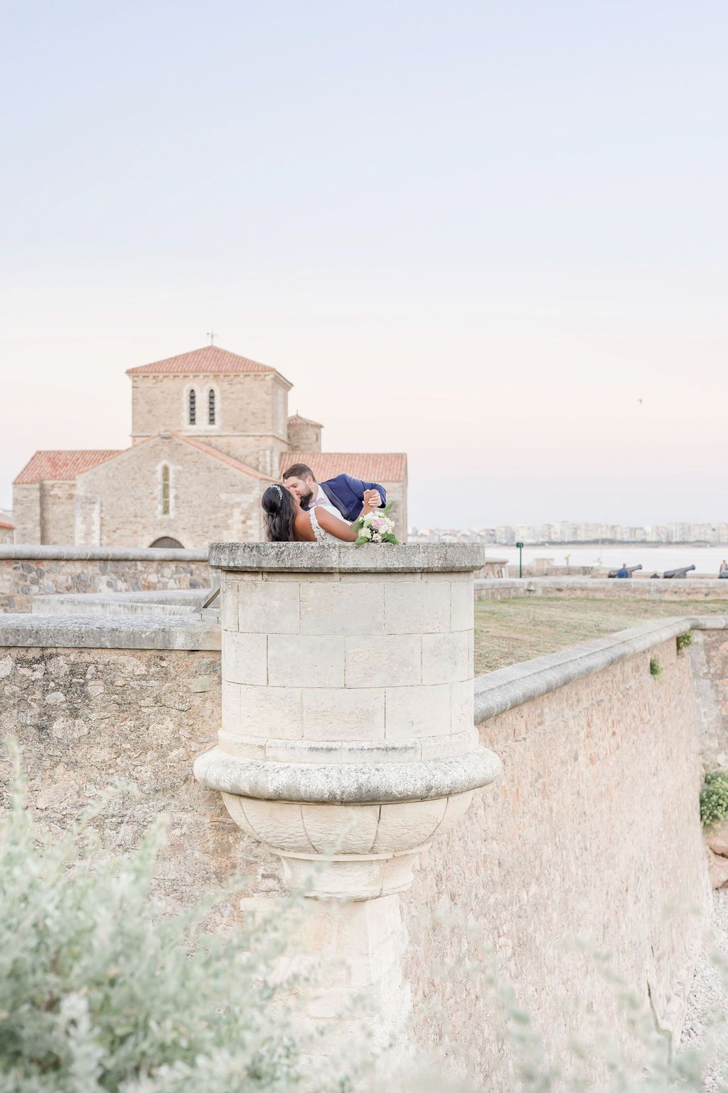 Couple embracing on stone pillar with historic church architecture in background.