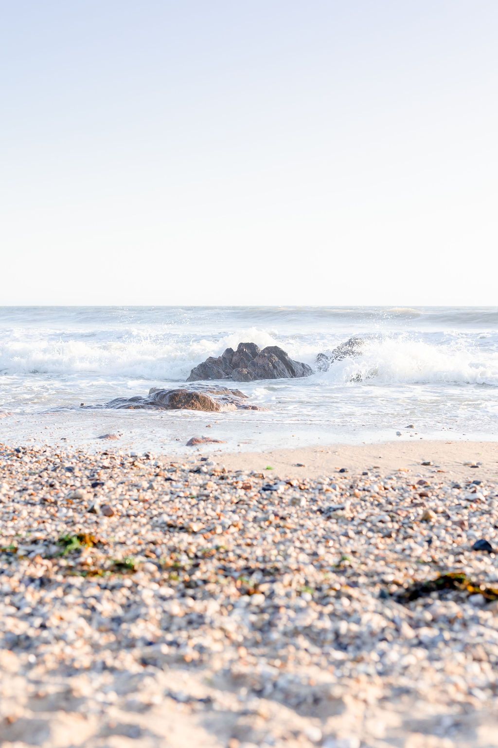 Serene ocean waves crash against rocks on a sunlit beach with scattered pebbles.