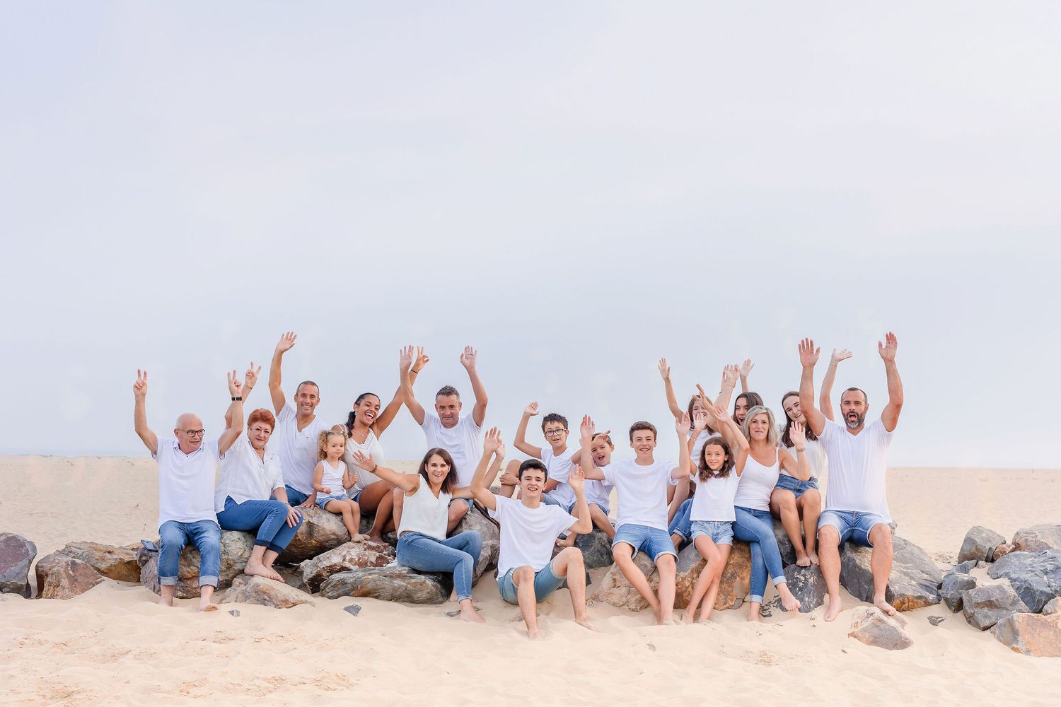 Extended family raises their arms joyfully while sitting together on a beach at sunset.