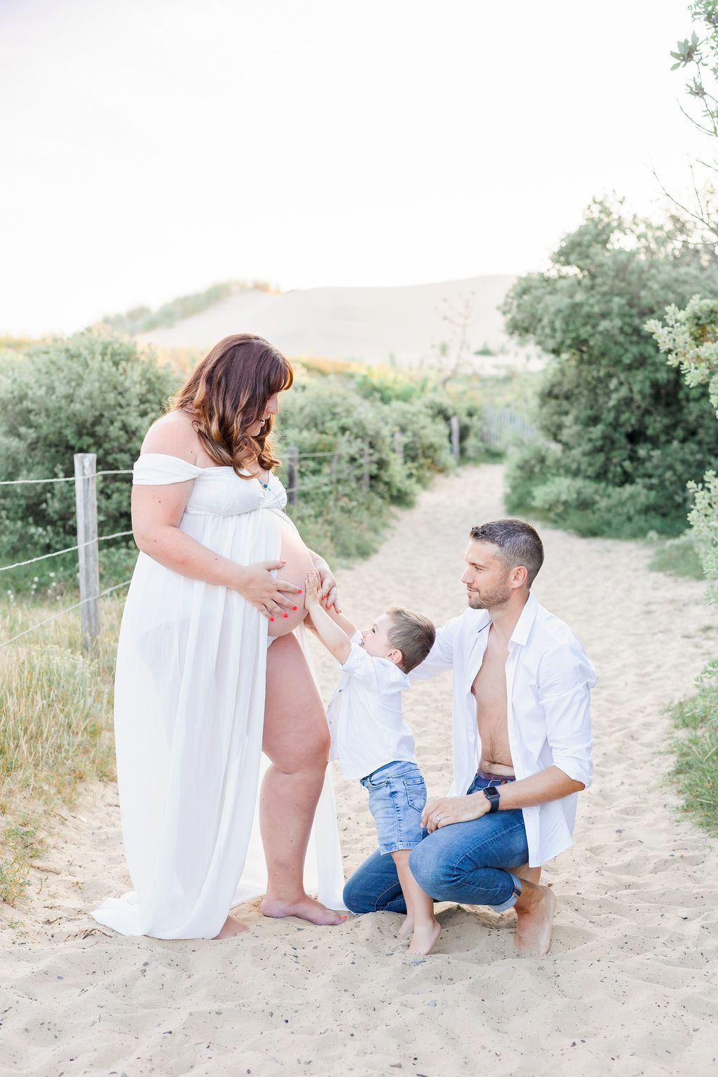 A heartwarming family maternity photo with a young child touching mom's baby bump on a sandy trail.