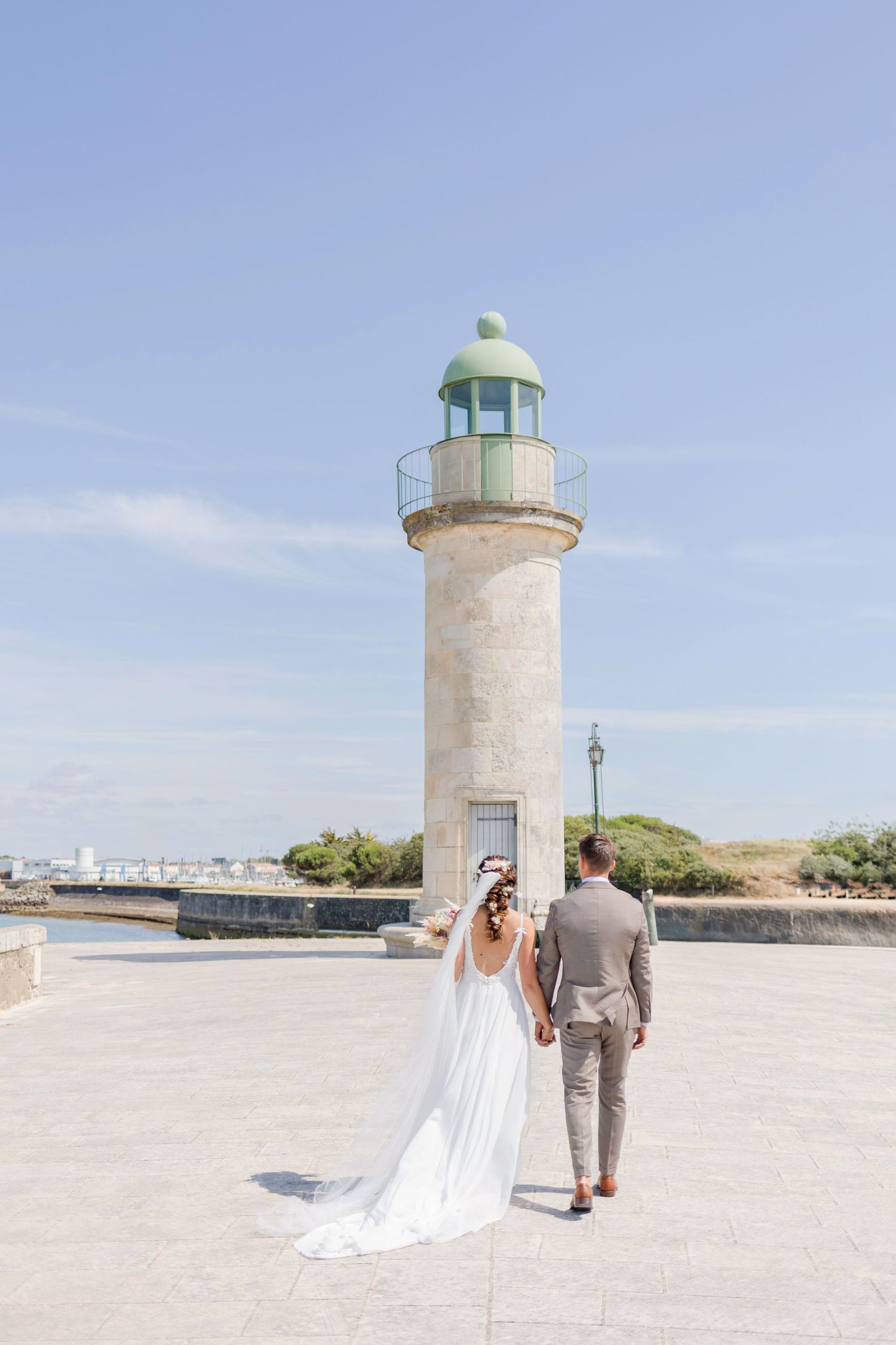 A couple in wedding attire walk towards a white lighthouse on a sunny day by the water.