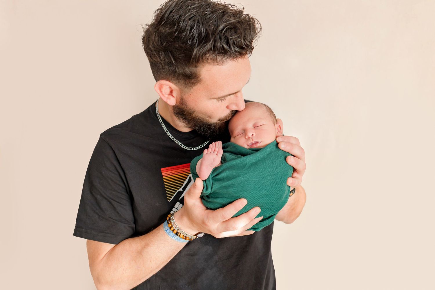 Parent tenderly holds sleeping newborn baby dressed in emerald green against neutral background.