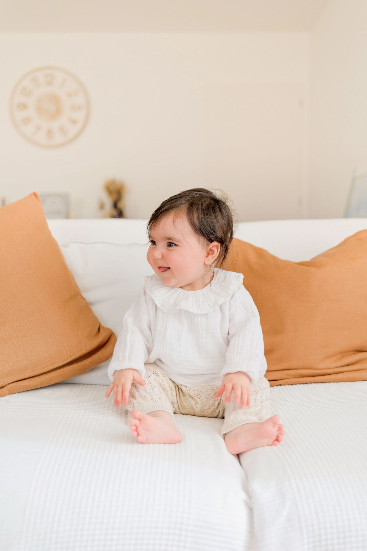 A small baby in white clothing sits on a white bed with orange accent pillows.