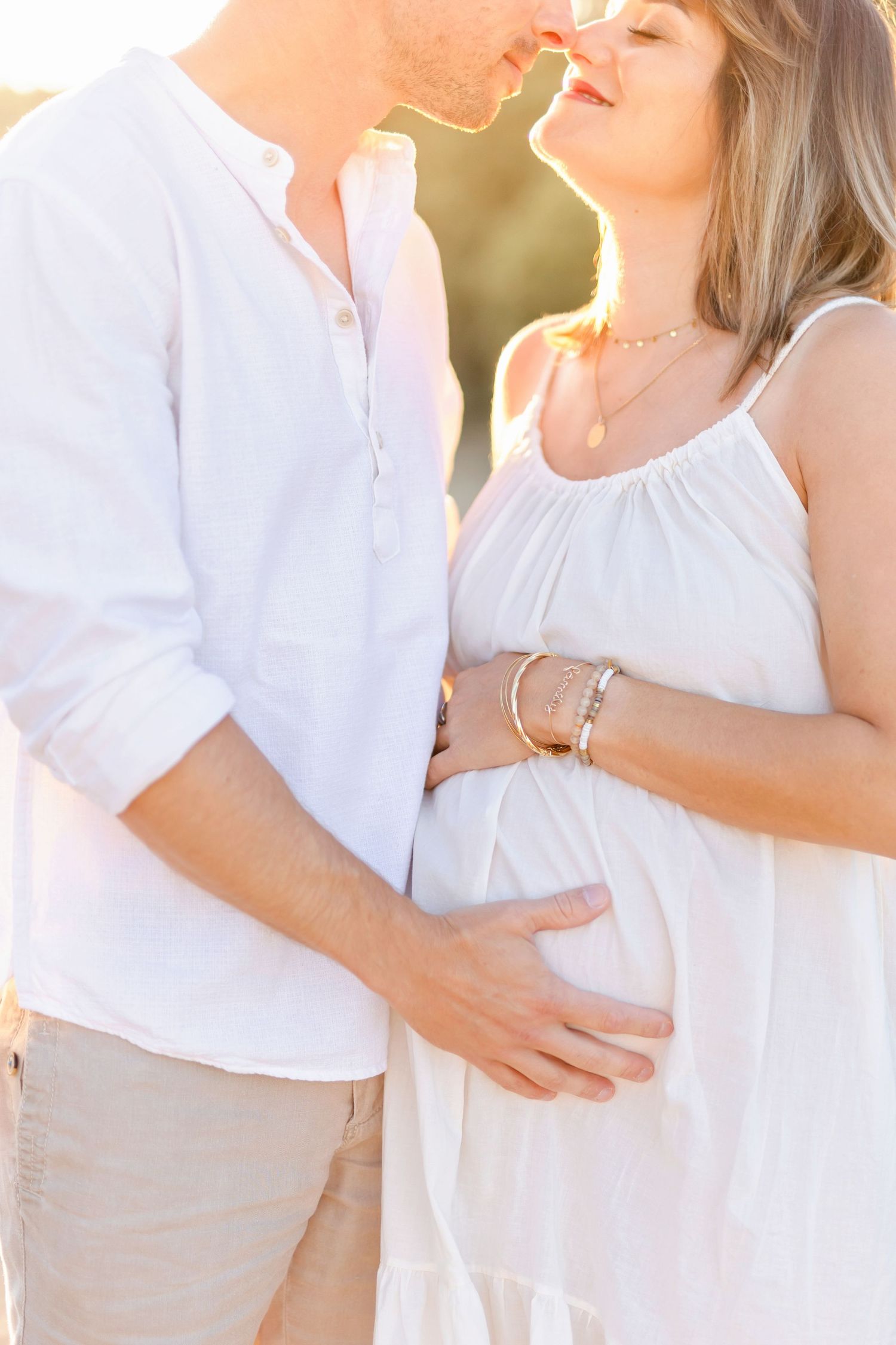 Sun-kissed maternity photo of couple in white clothing sharing an intimate moment outdoors.