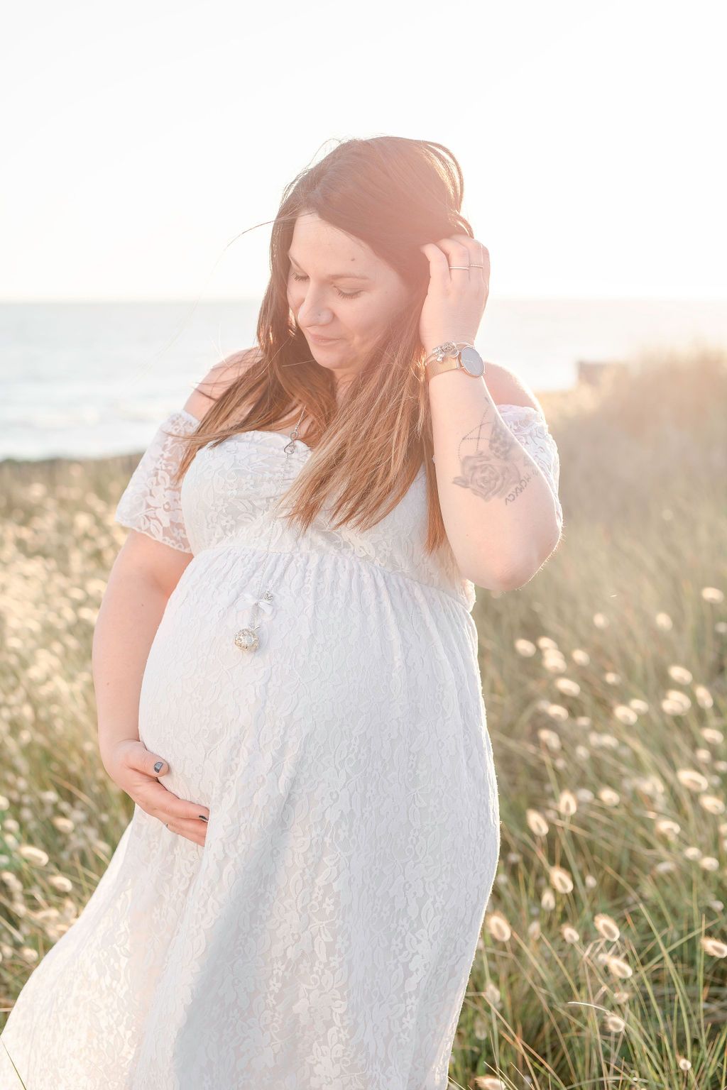Pregnant woman in white lace dress touches her hair during sunset beach photoshoot.