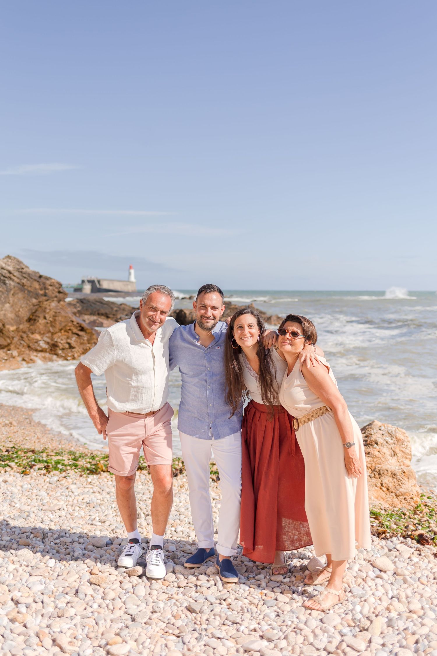 Four people stand together smiling on a rocky coastal beach with a lighthouse visible in the distance.