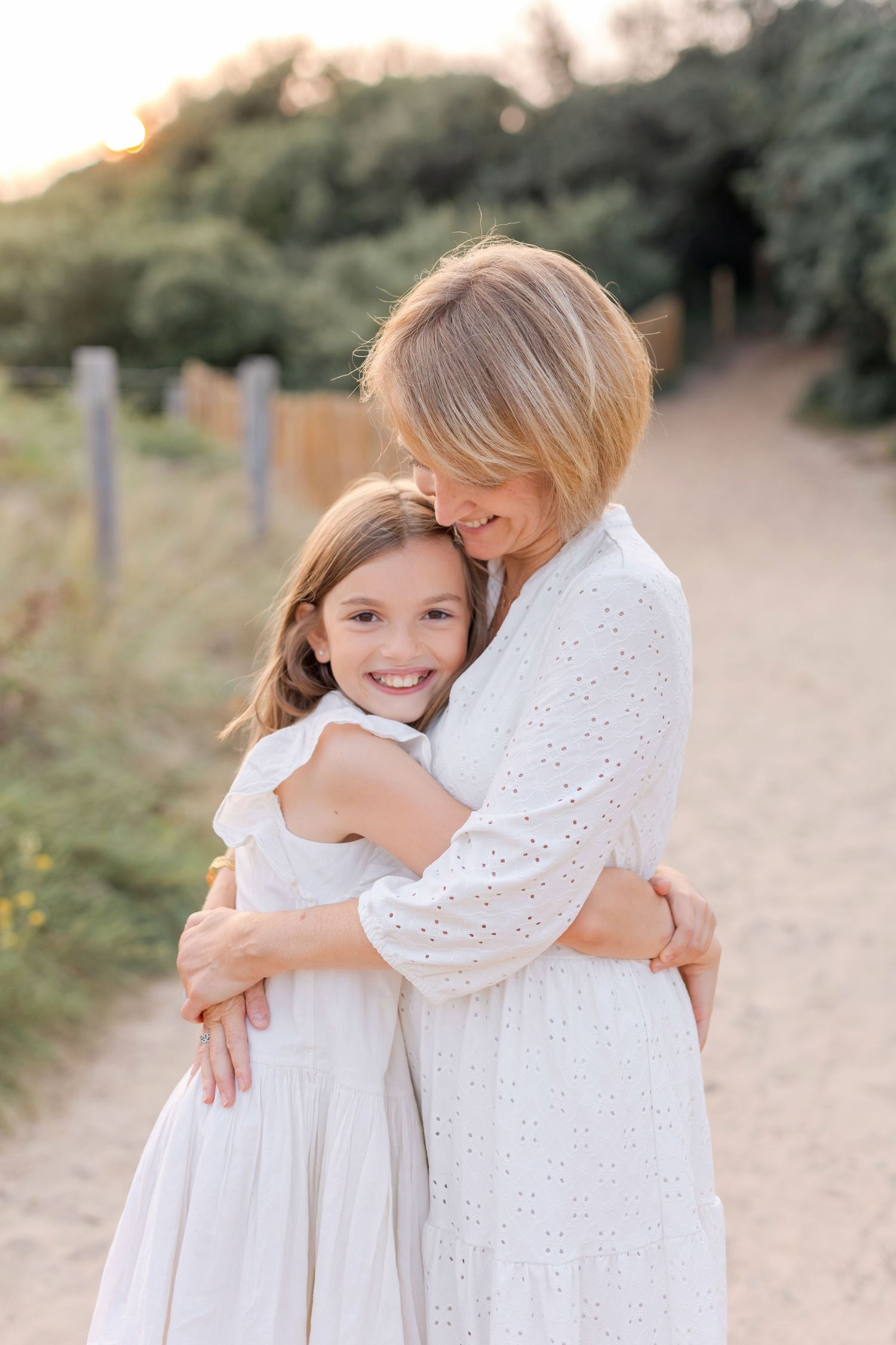 Parent and child in white dresses sharing tender embrace on nature path at sunset.