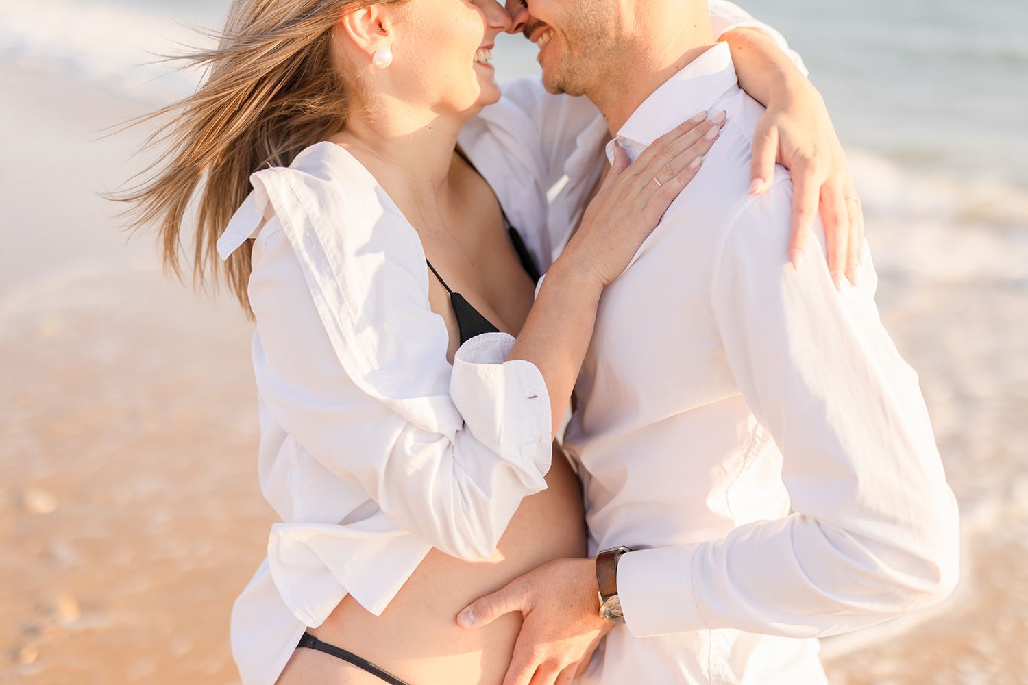 A romantic embrace between two people wearing white outfits standing along a sunny beach shoreline.