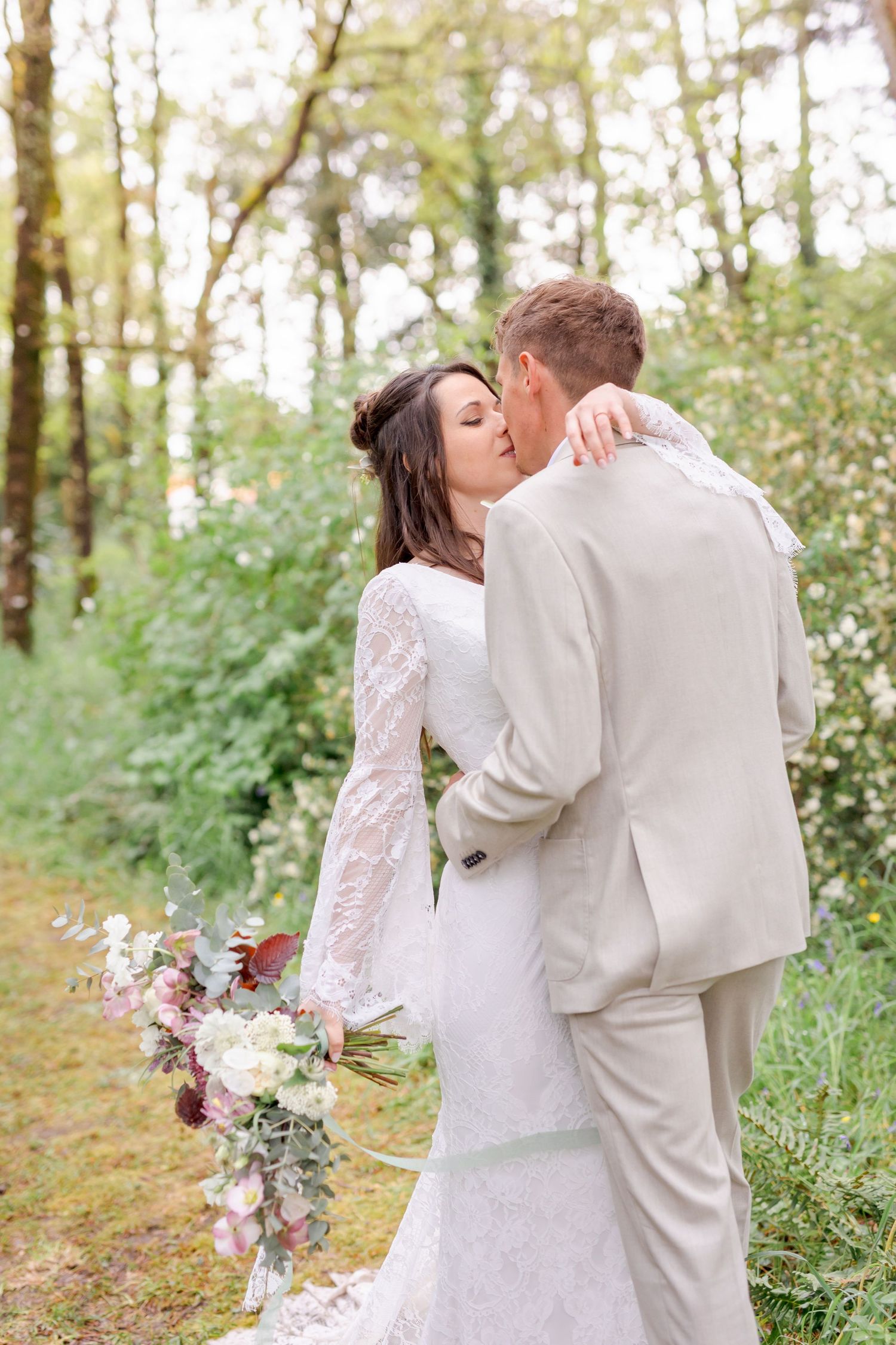 Wedding couple embraces on a sunlit forest path with bride holding a pink and white bouquet.