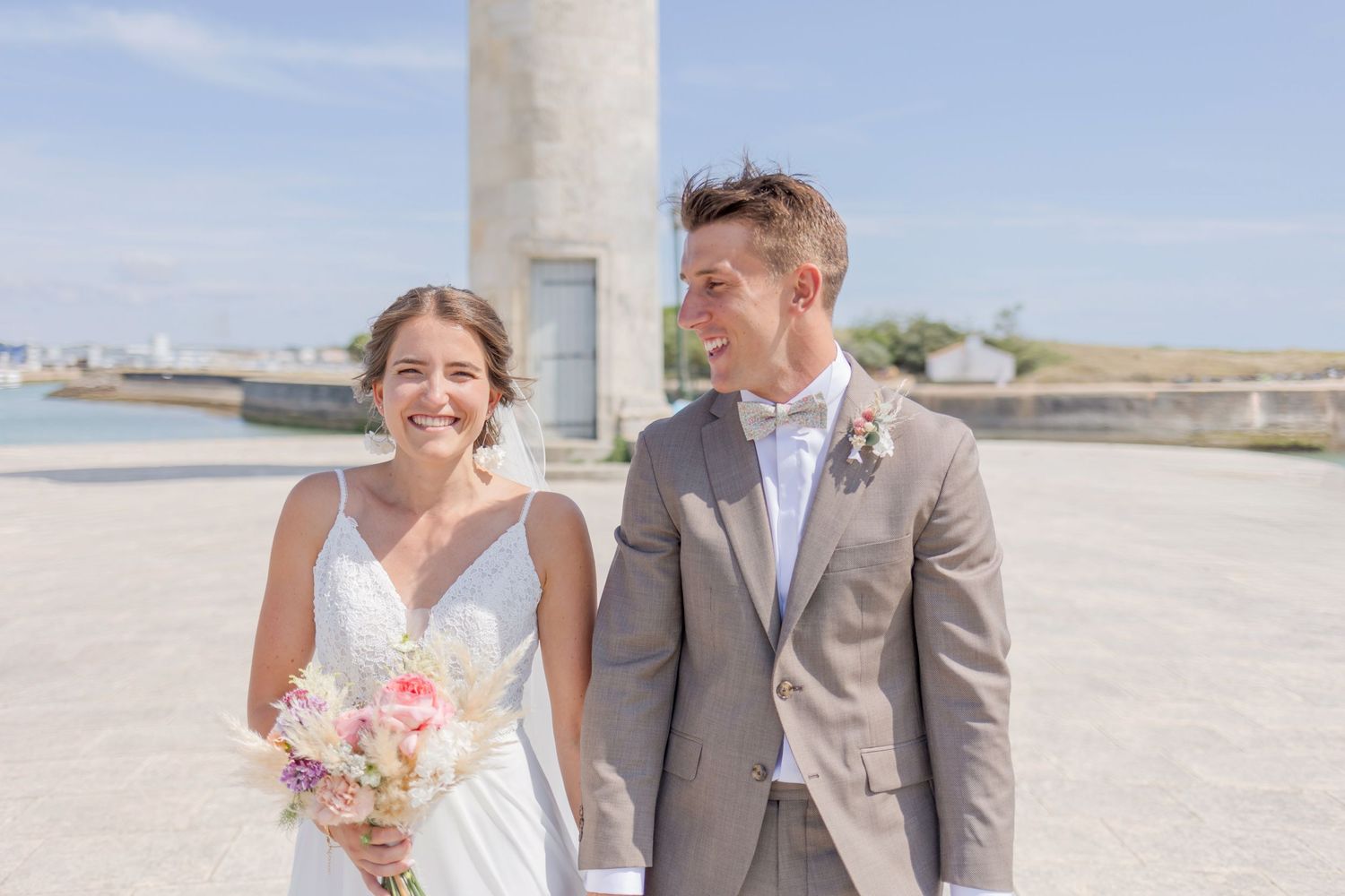 Happy couple in wedding attire smiling near lighthouse with pink bouquet.