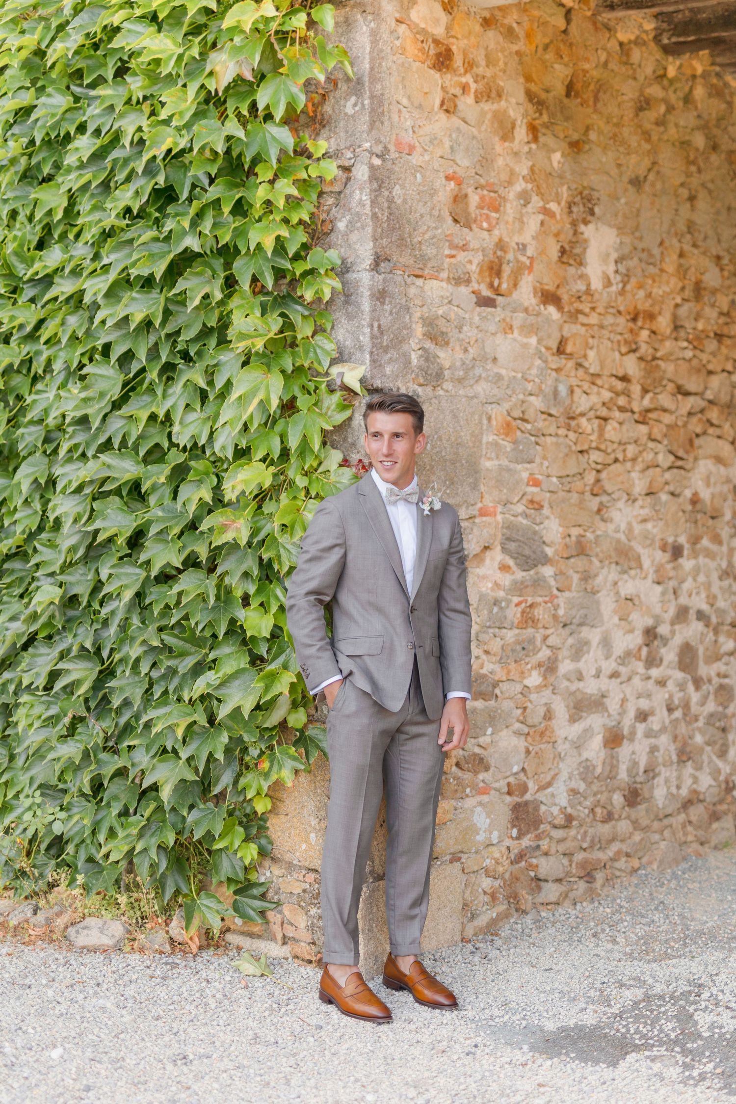 Groom in light grey suit and brown shoes poses against ivy-covered stone wall.