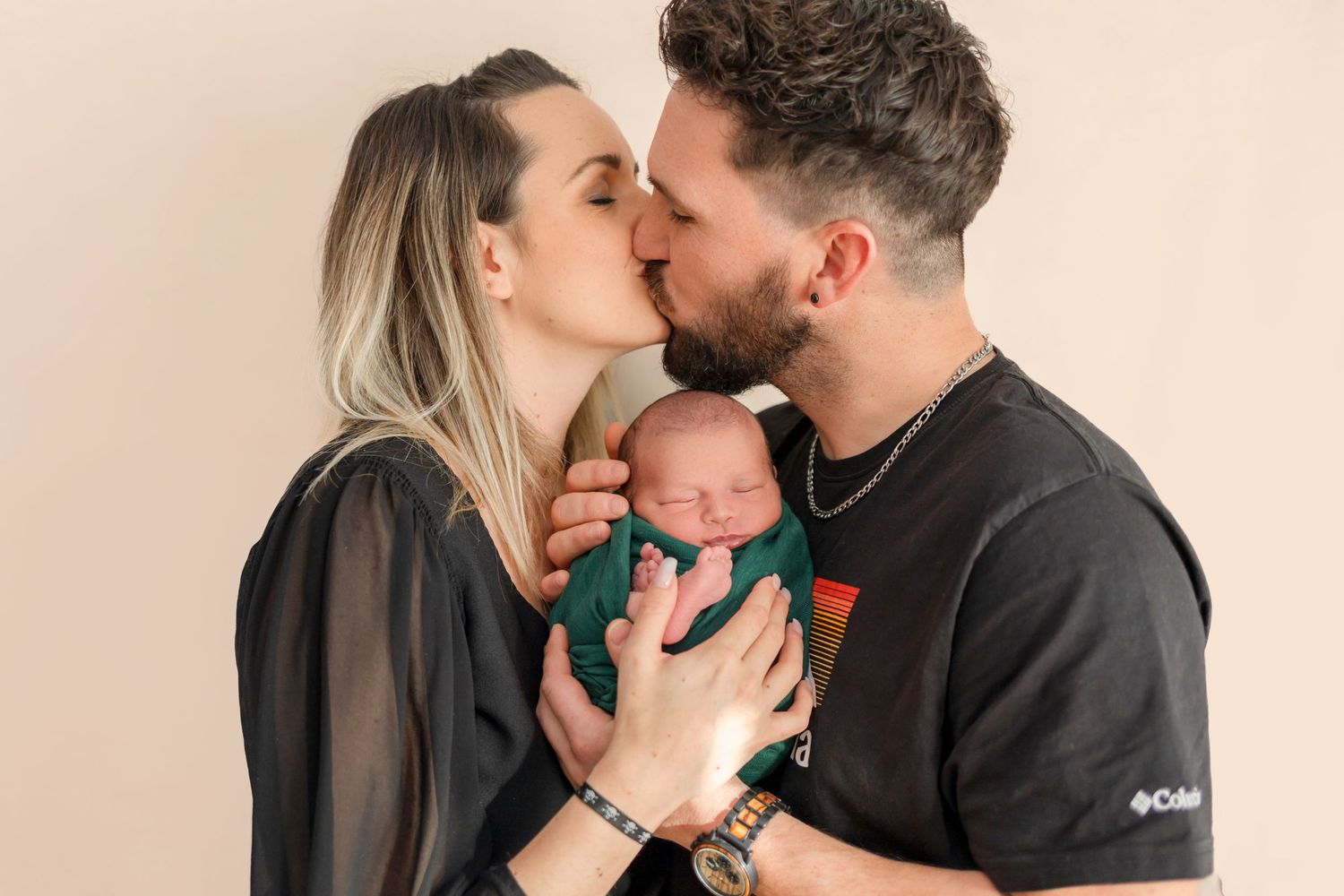 Parents kiss while holding their peacefully sleeping newborn between them.