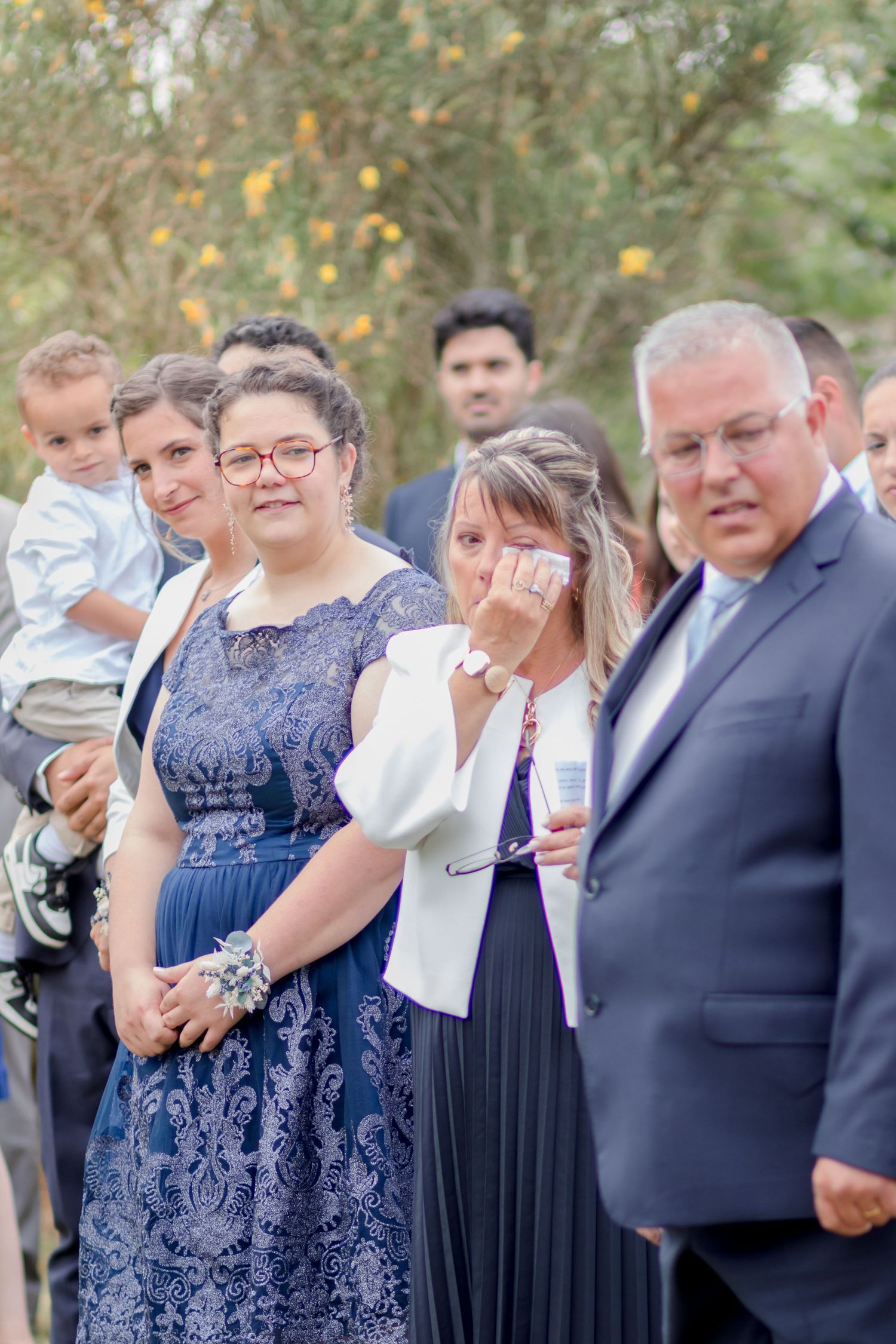 Wedding guests show emotional reactions during outdoor ceremony with lush greenery in background.