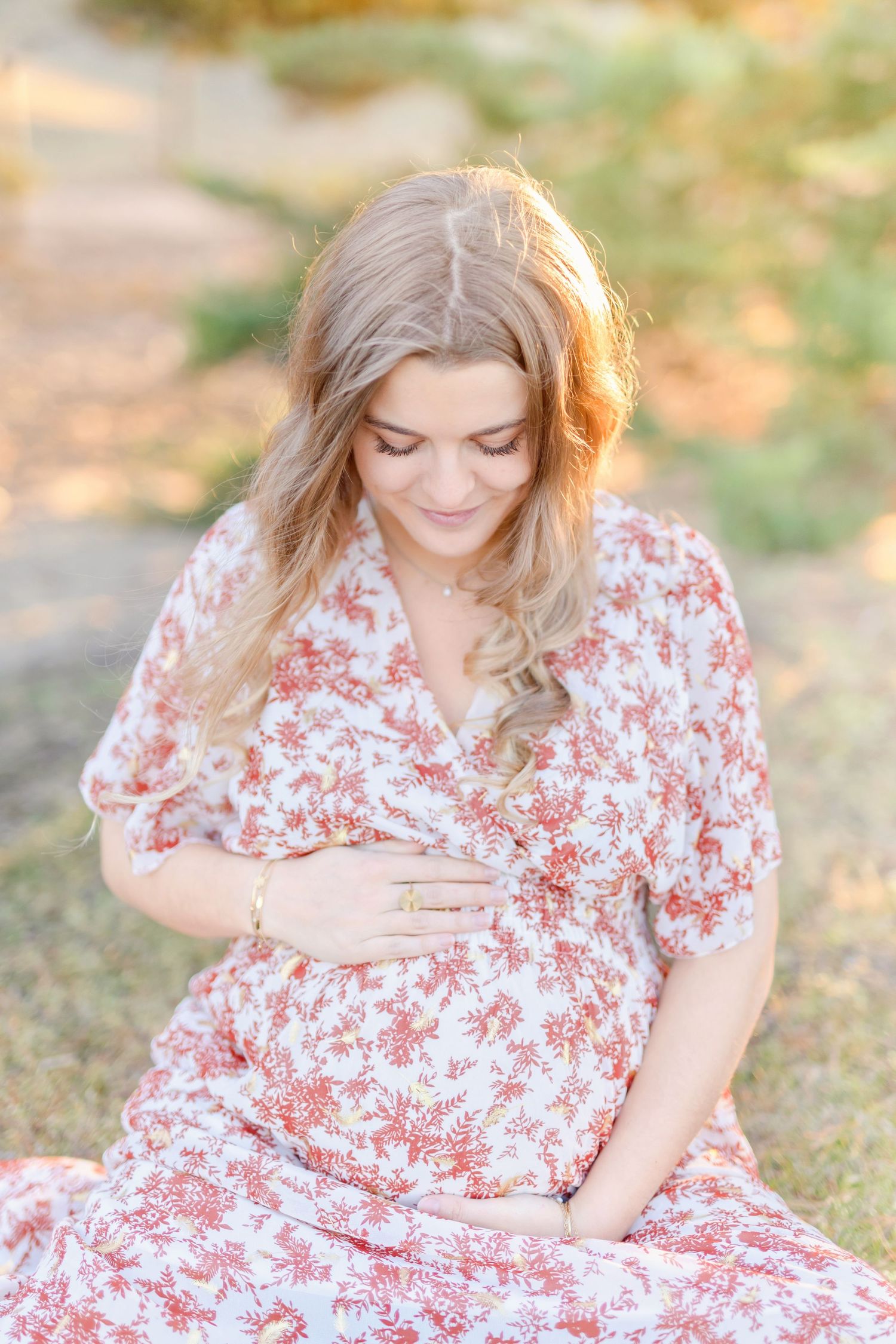 Woman in floral dress gently touching her baby bump in soft sunlight.