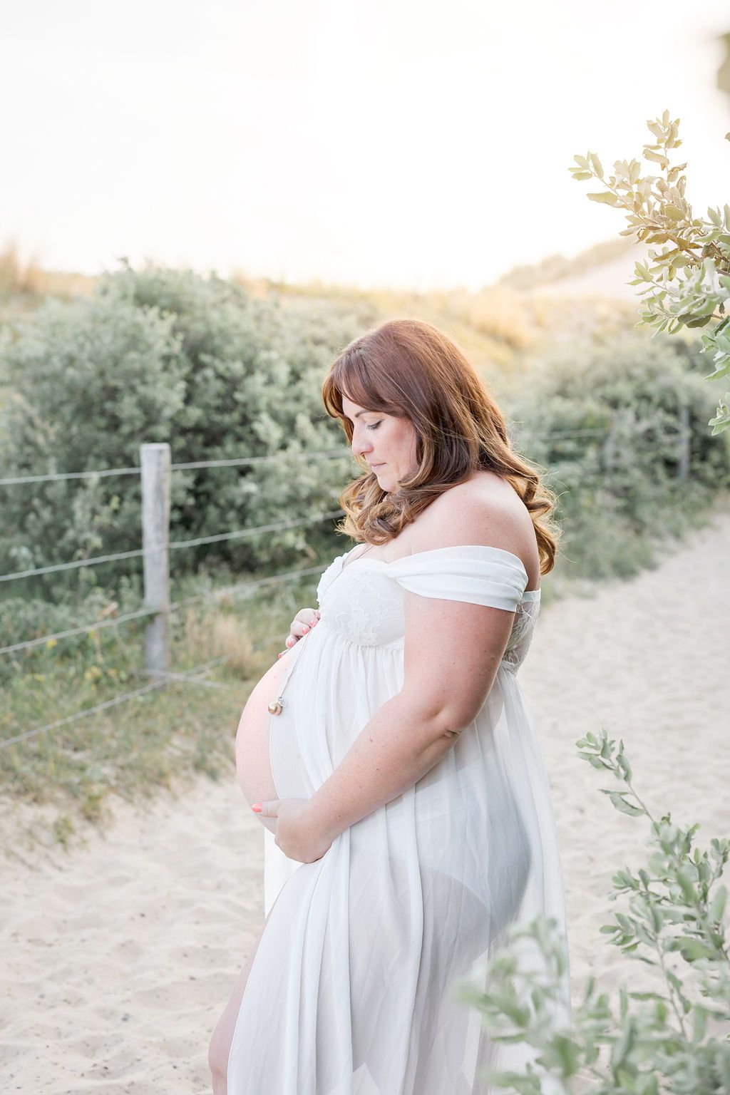 An elegant maternity portrait of an expectant mother in a flowing white dress against a natural landscape.