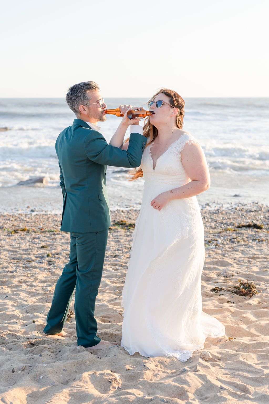 Couple in wedding attire enjoying beverages while sitting on sandy beach by the ocean.
