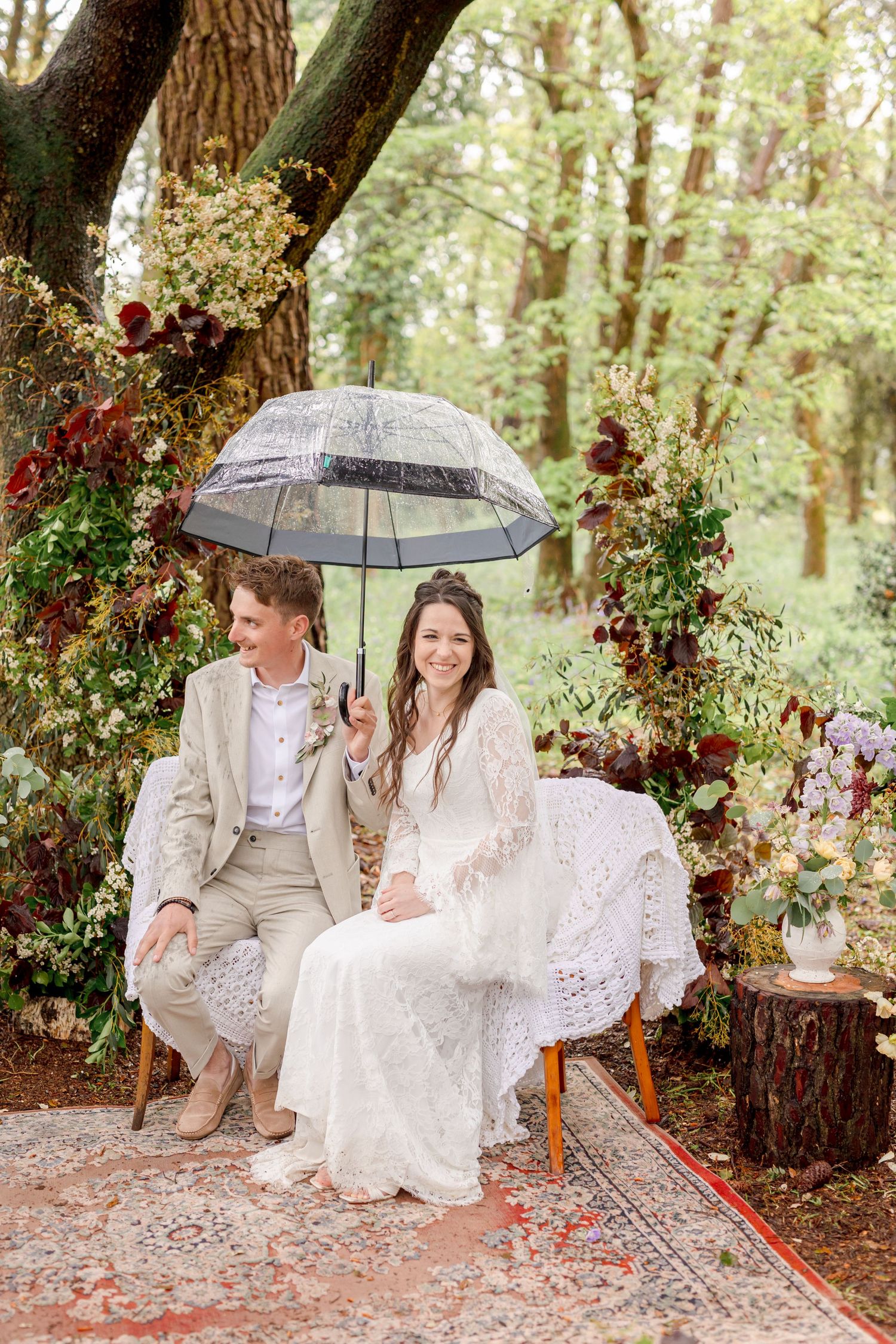 Couple poses together on decorative rug under umbrella in woodland setting.