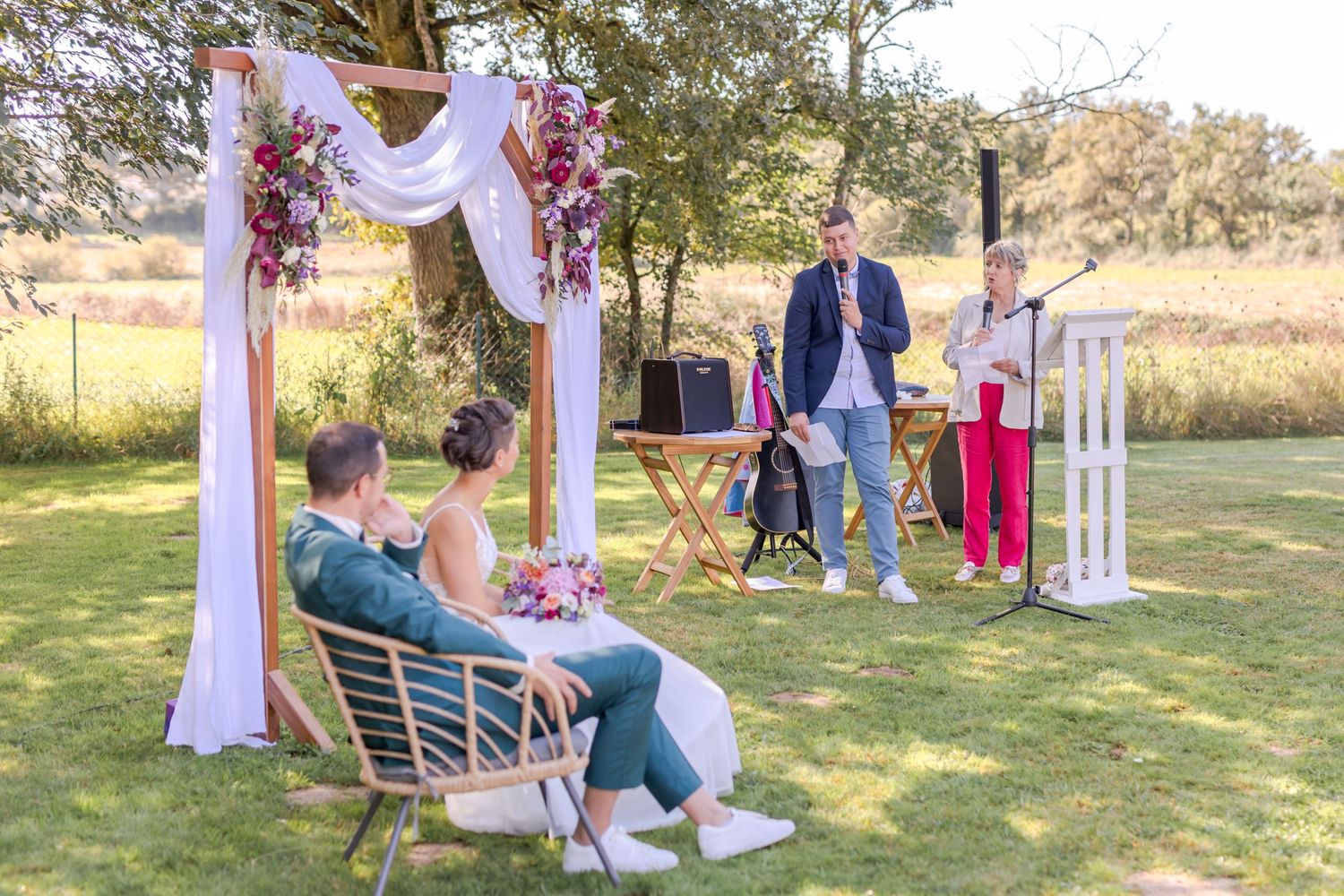 Casual outdoor wedding setup with white arch and scattered seating on lawn.