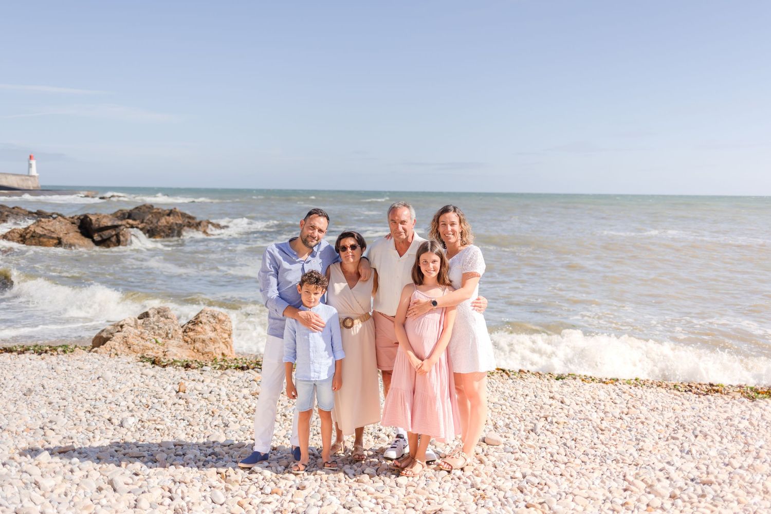 A family group stands together on a pebble beach with ocean waves and a lighthouse in the distance.