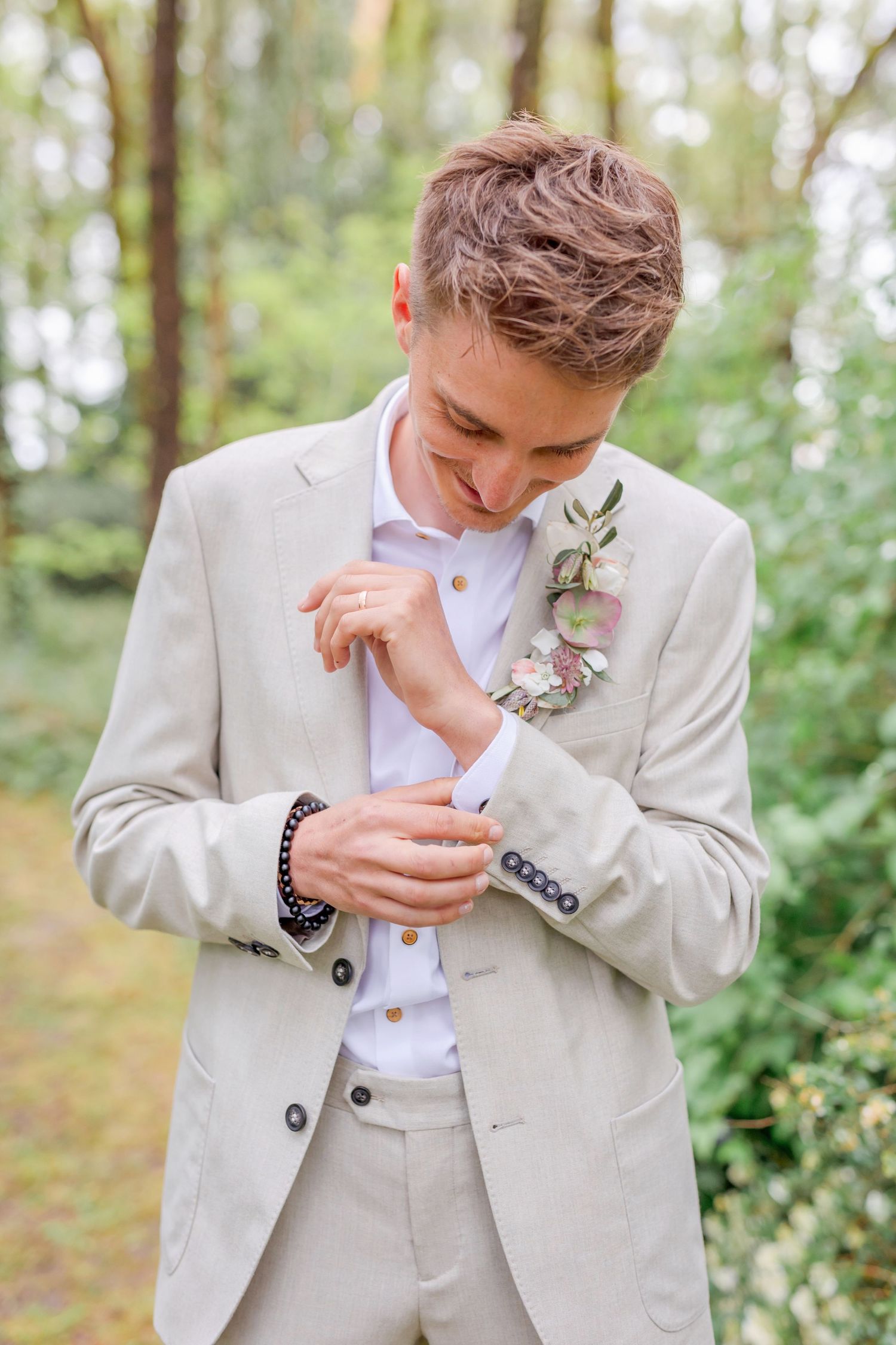Groom in a light beige suit adjusts his cufflink while wearing a delicate floral boutonniere.