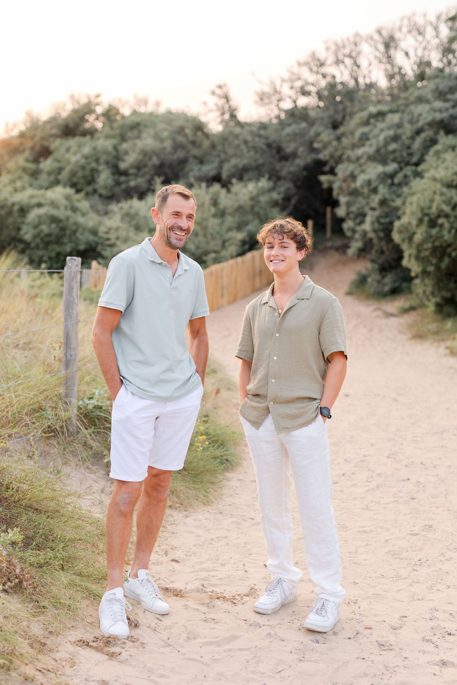 Two people in casual summer outfits standing on a dirt path surrounded by trees.