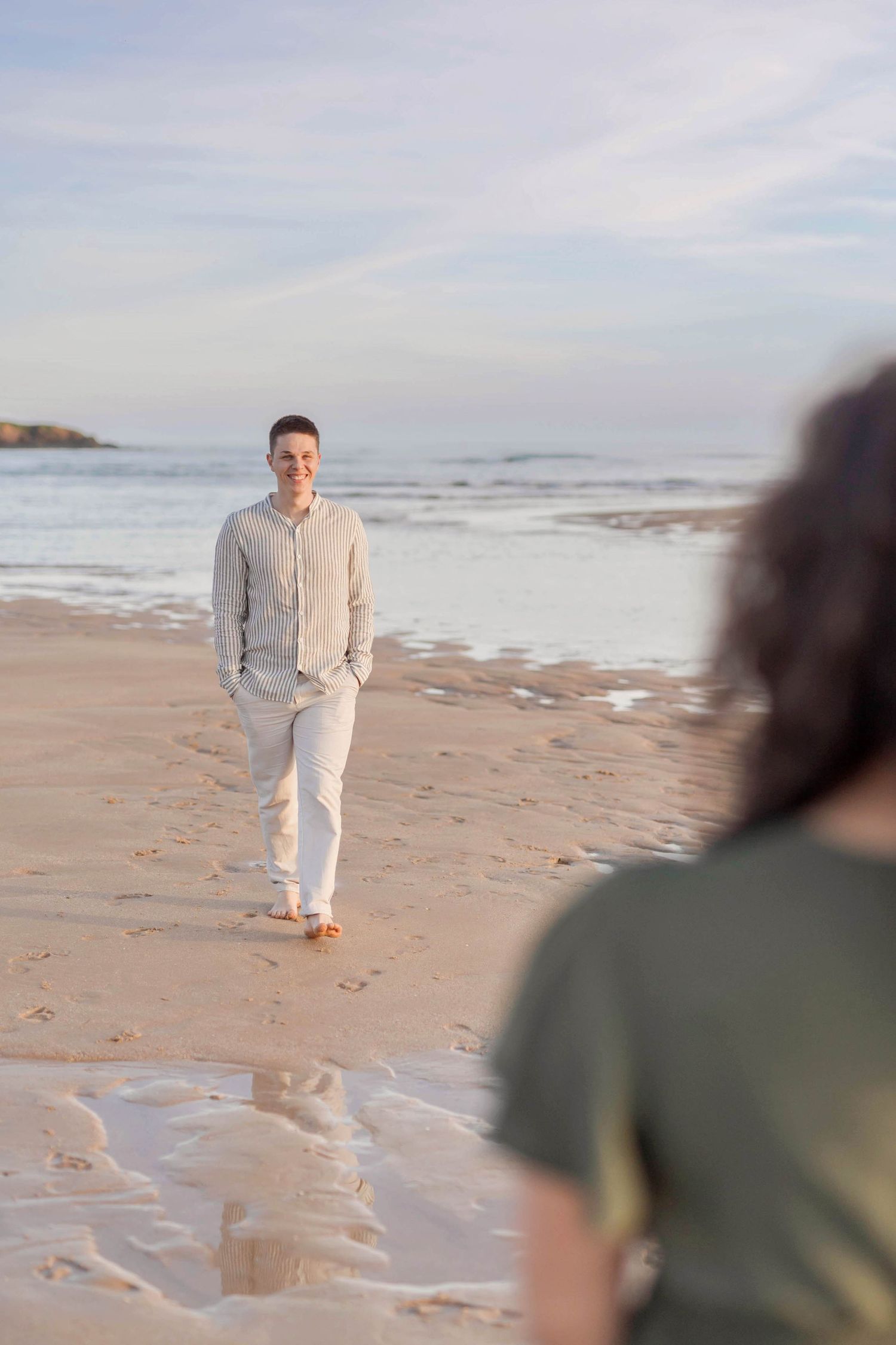 A person in white clothing stands on a beach at sunset with waves in the background.