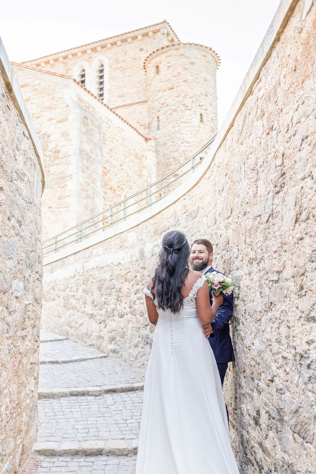 Wedding couple poses on stone steps against rustic Mediterranean wall.