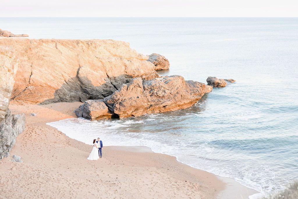 Newlyweds walk along a scenic rocky beach with towering cliffs and blue ocean waters.