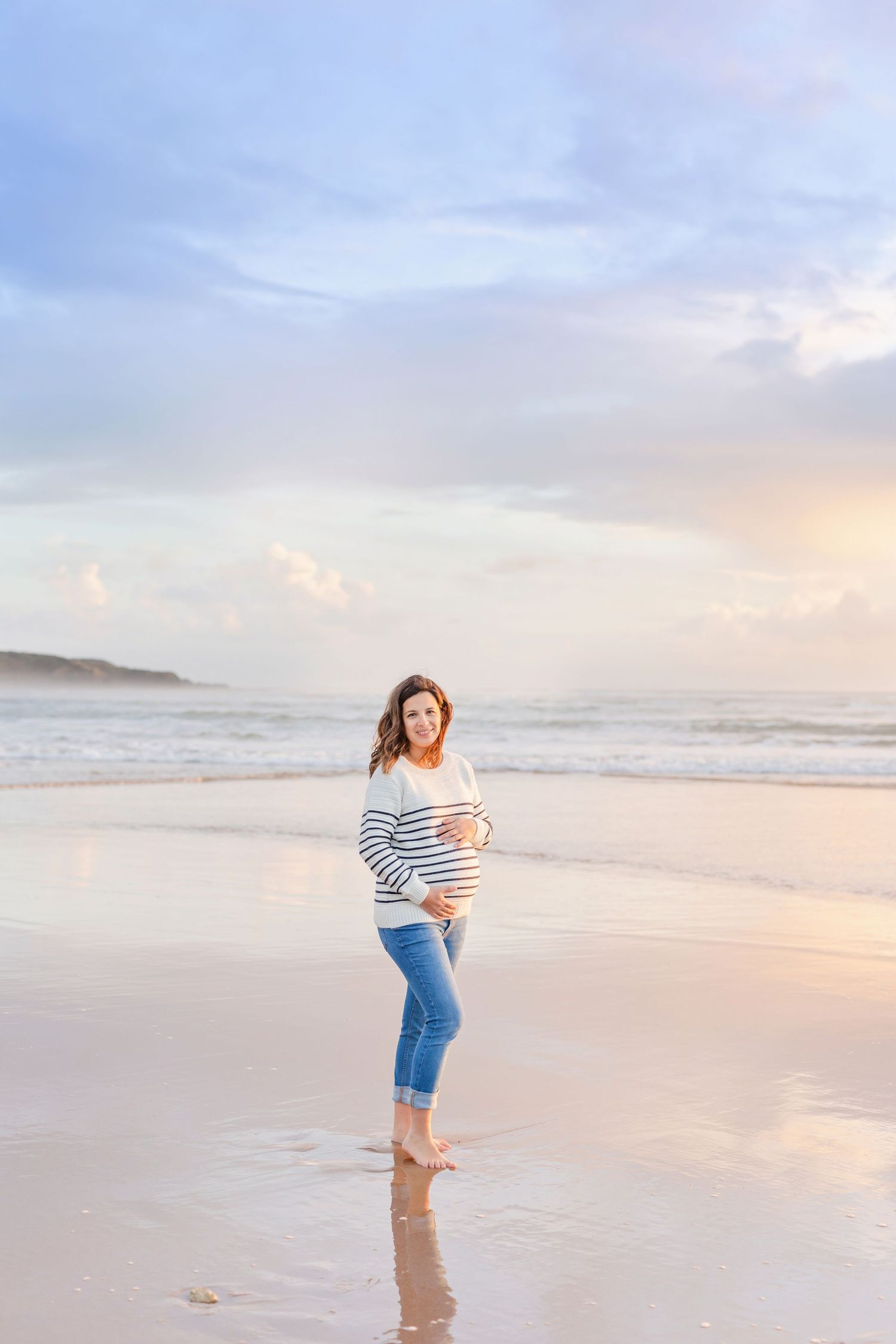 Expecting mother walking along empty beach shoreline at golden hour wearing striped sweater and jeans.