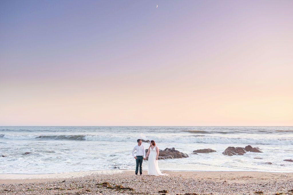 A couple in wedding attire stands on beach with waves and rocks in background.
