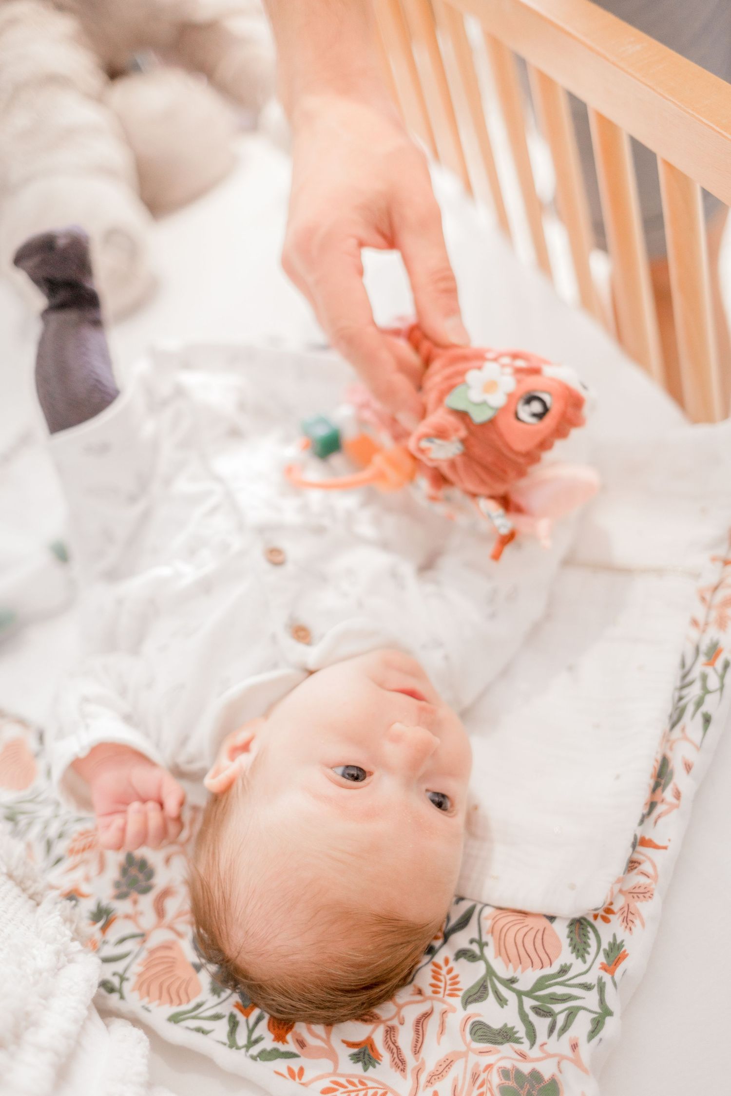 Orange plush toy rests beside infant on floral bedding in crib.