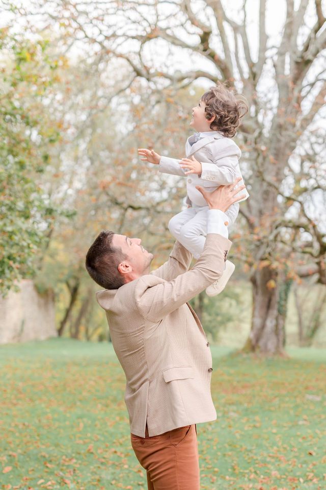 Playful outdoor portrait with light beige suit lifting small figure in white against bare autumn trees.