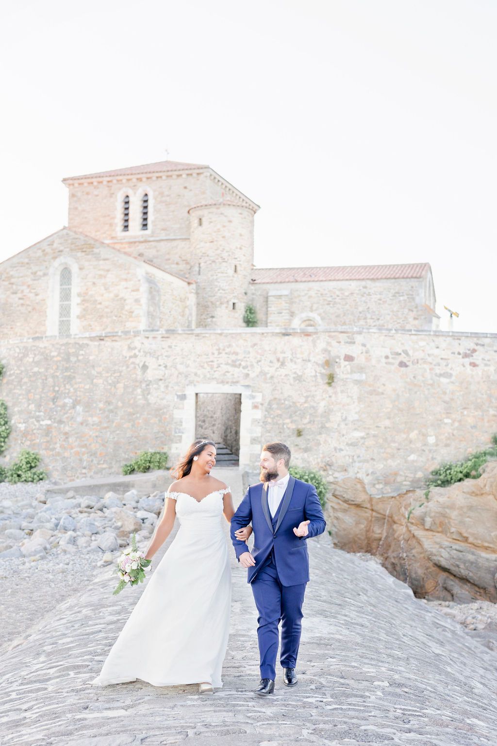 Wedding couple poses on cobblestone path near historic stone building.