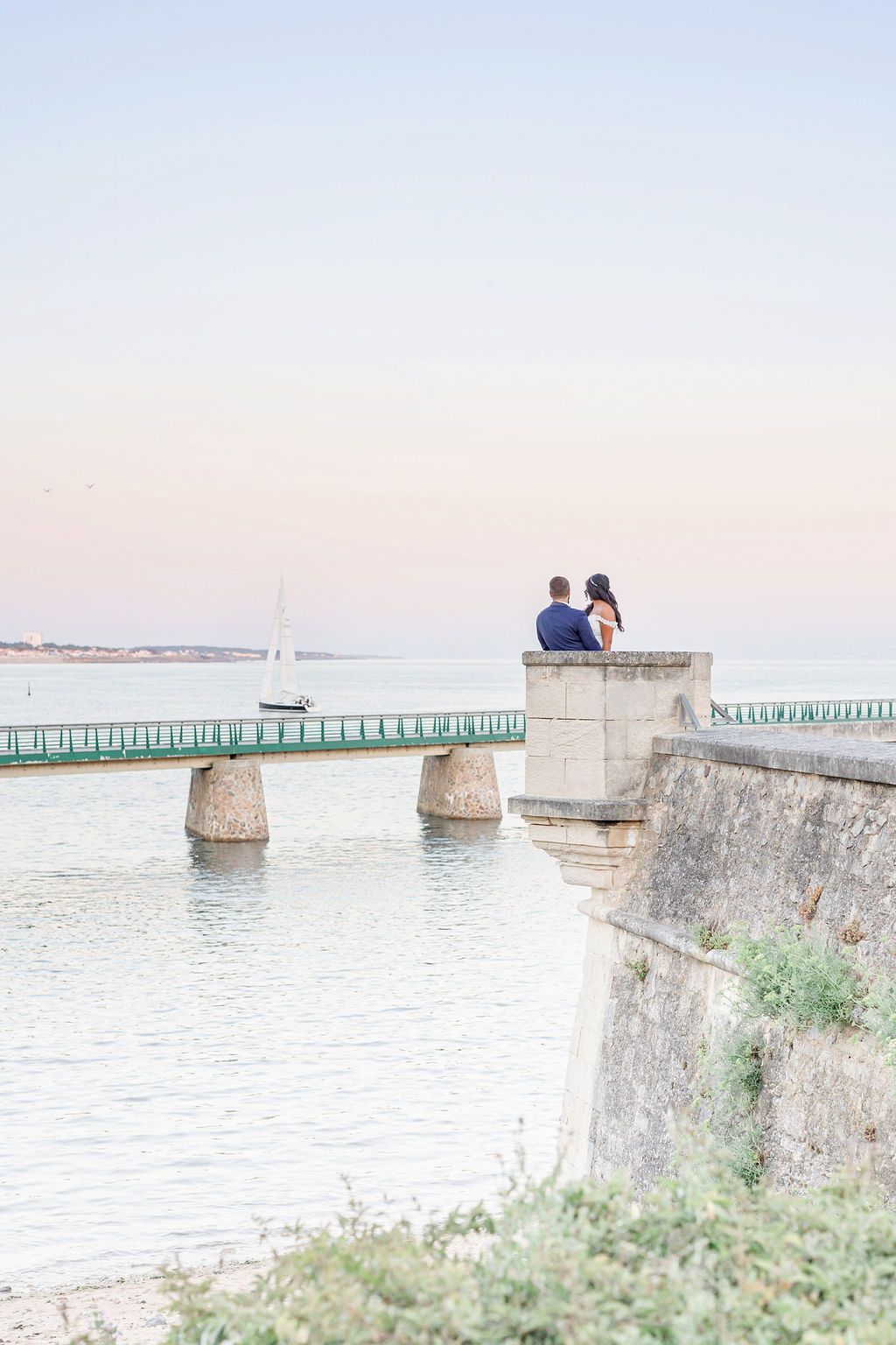 Wedding couple sits on coastal wall with sailboat and bridge visible in distance.