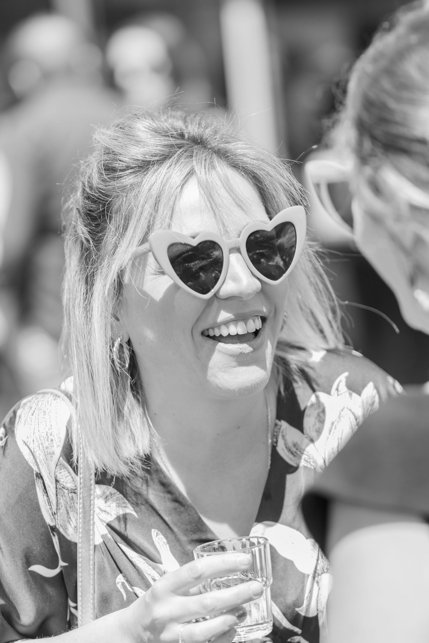 Woman wearing heart-shaped sunglasses smiles brightly at outdoor event.