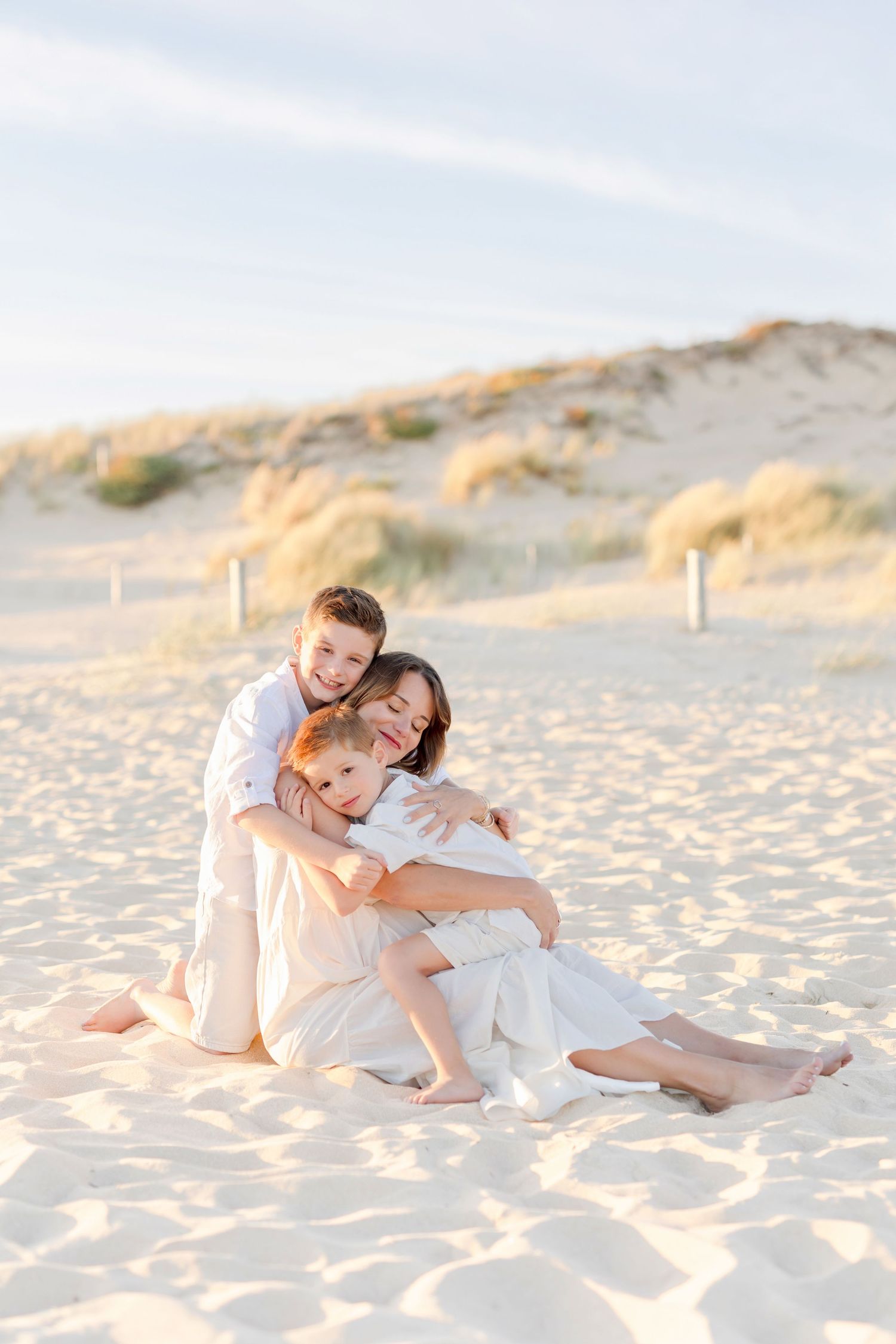 Family group hug moment captured on sandy beach with dunes backdrop.