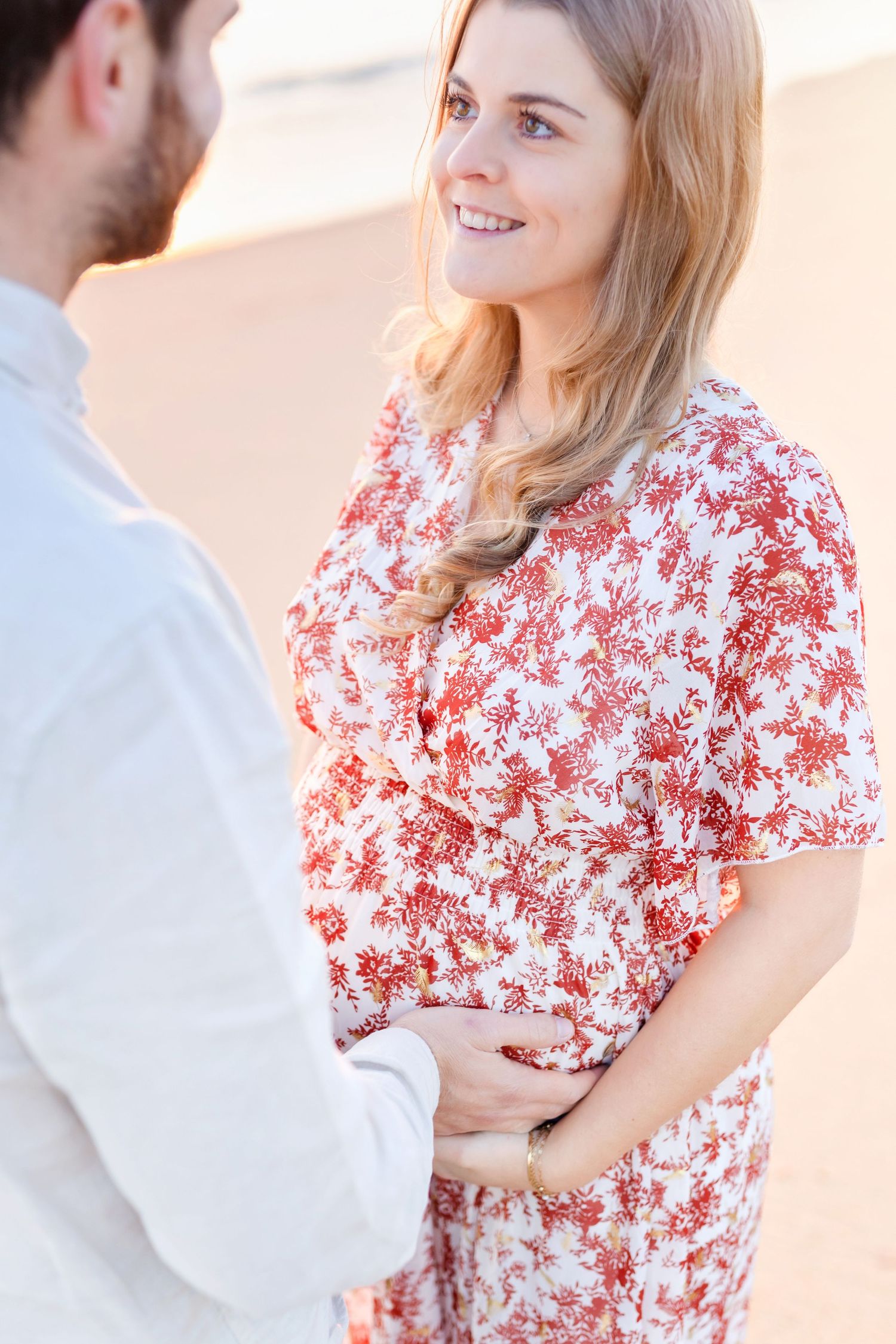 Couple in romantic beach photoshoot with woman wearing red and white floral dress.
