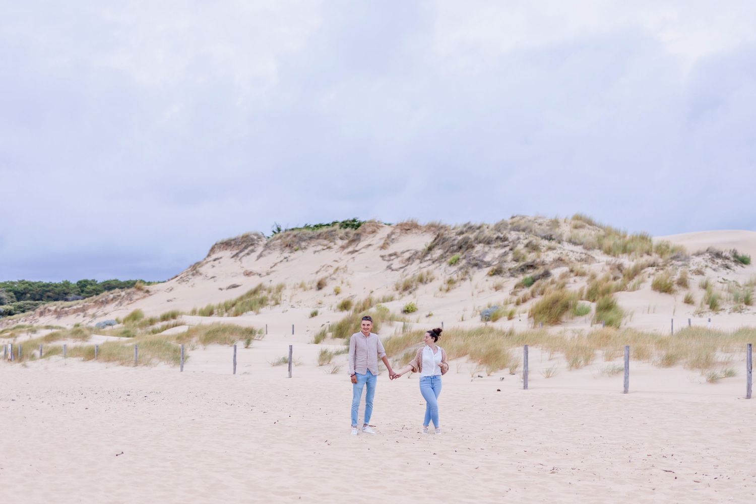 Two people walking together on sandy path through coastal dunes under cloudy sky.
