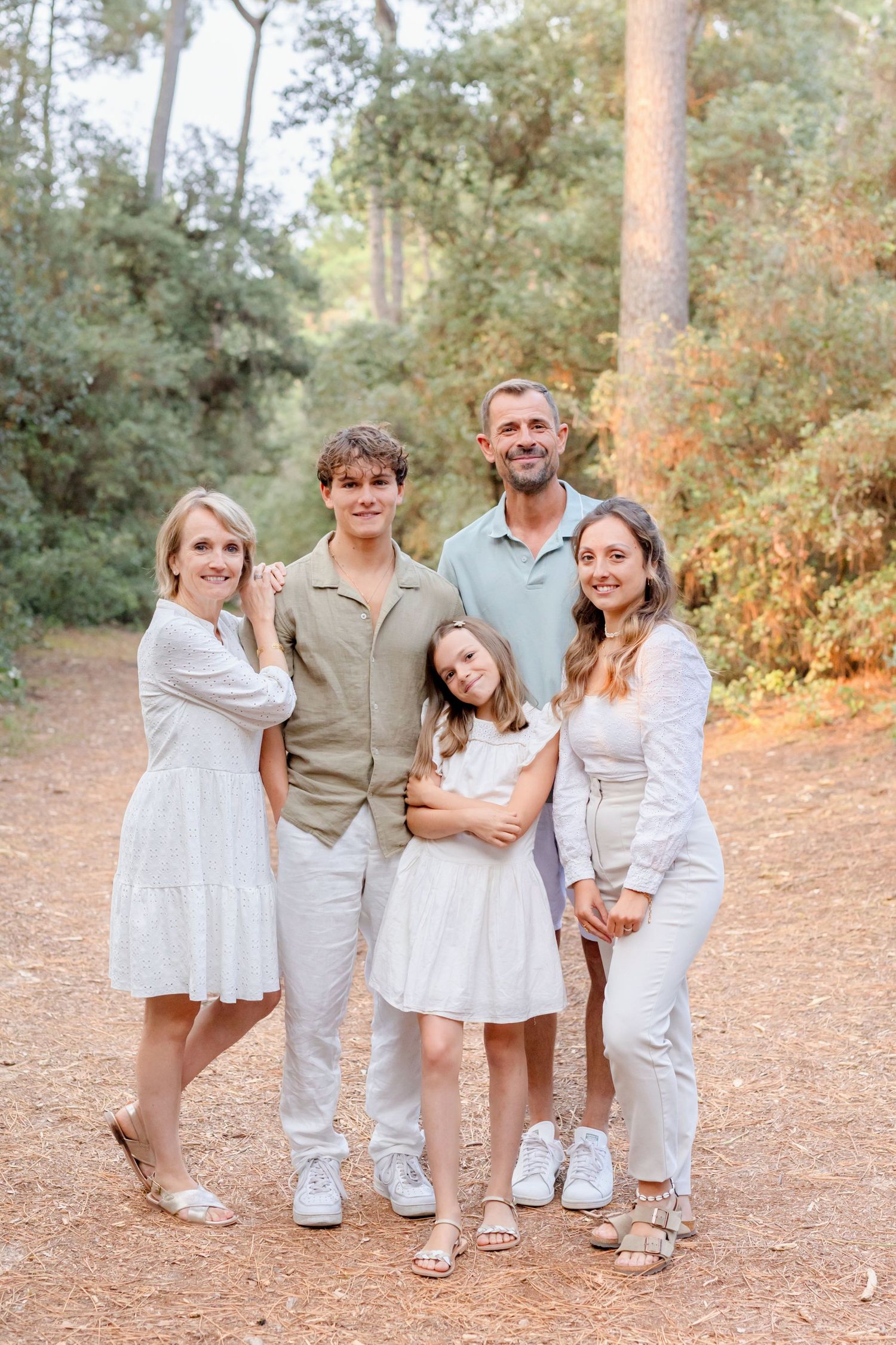 A family group in matching light colored outfits poses casually together on a wooded path.