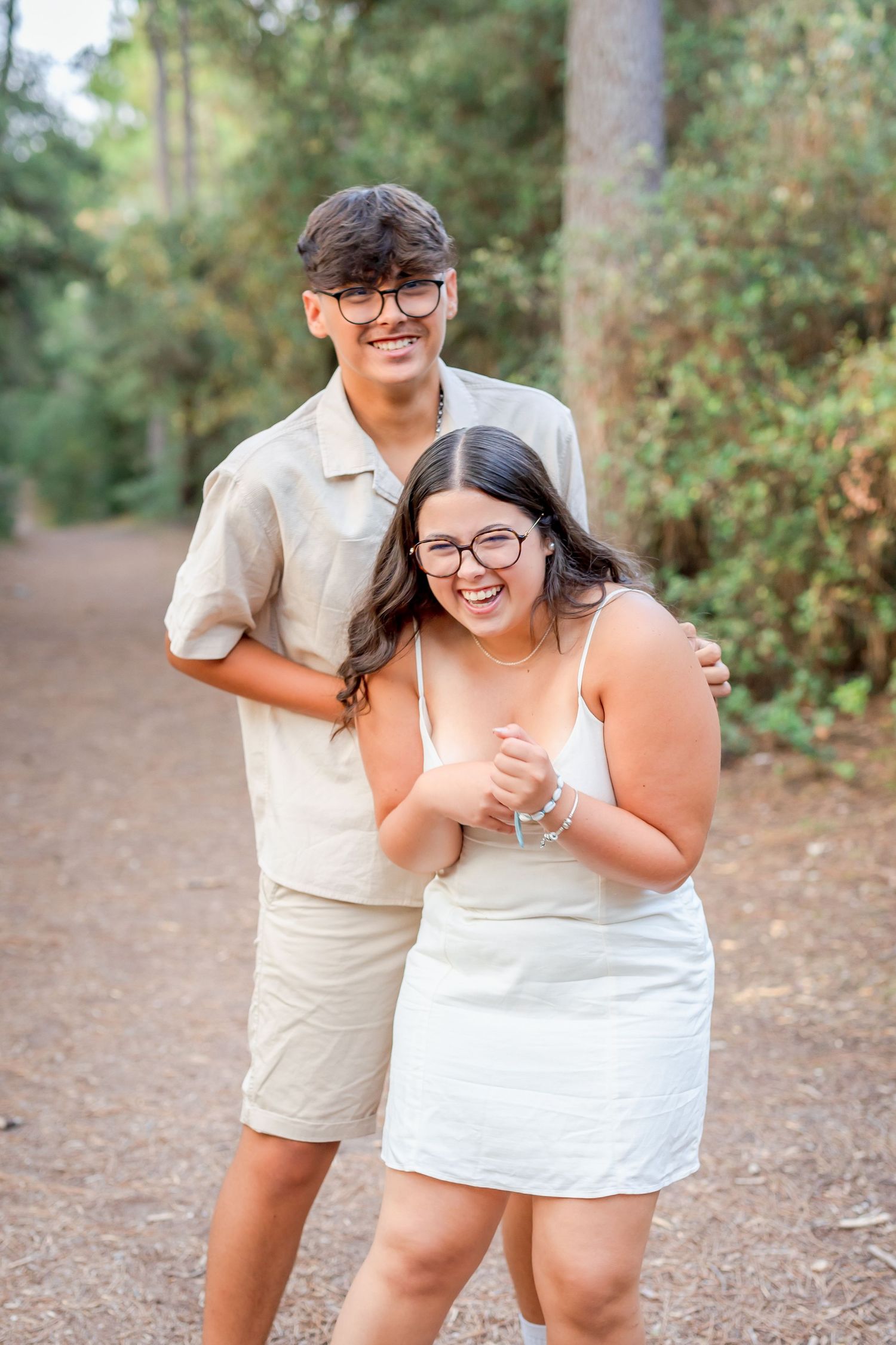 A joyful couple in coordinating cream-colored outfits laughing together on a wooded path.