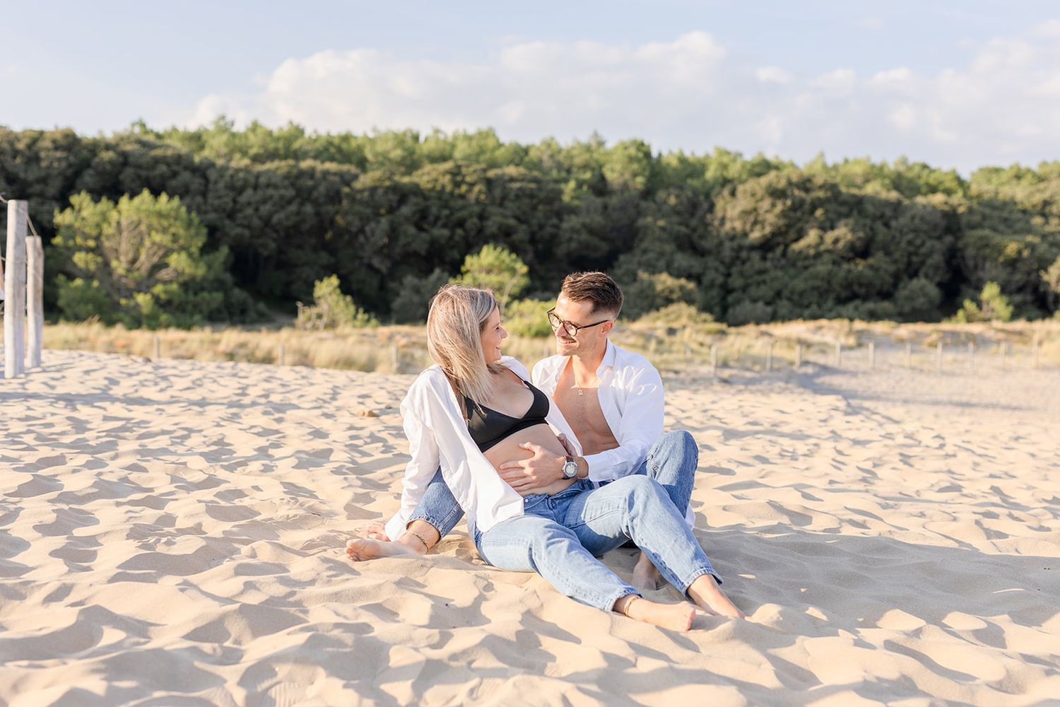 Two people sitting together on a sandy beach with trees and blue sky in the background.