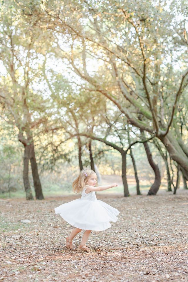 Young child in white dress twirling joyfully among trees in dreamy sunlit forest.