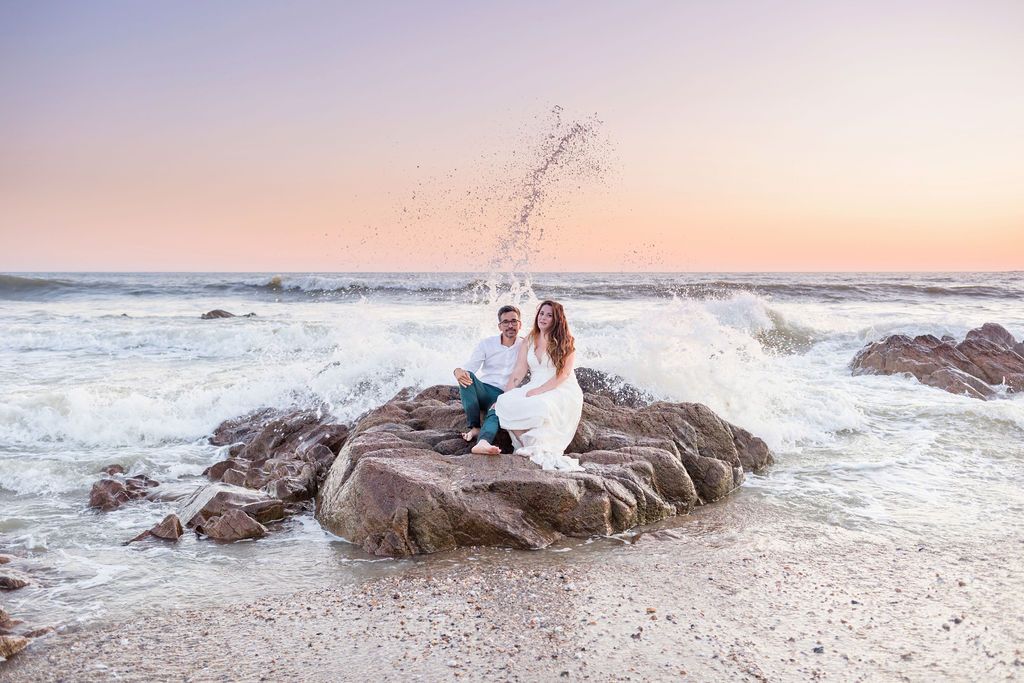 A couple sits on rocky beach outcrop with waves splashing behind them at sunset.