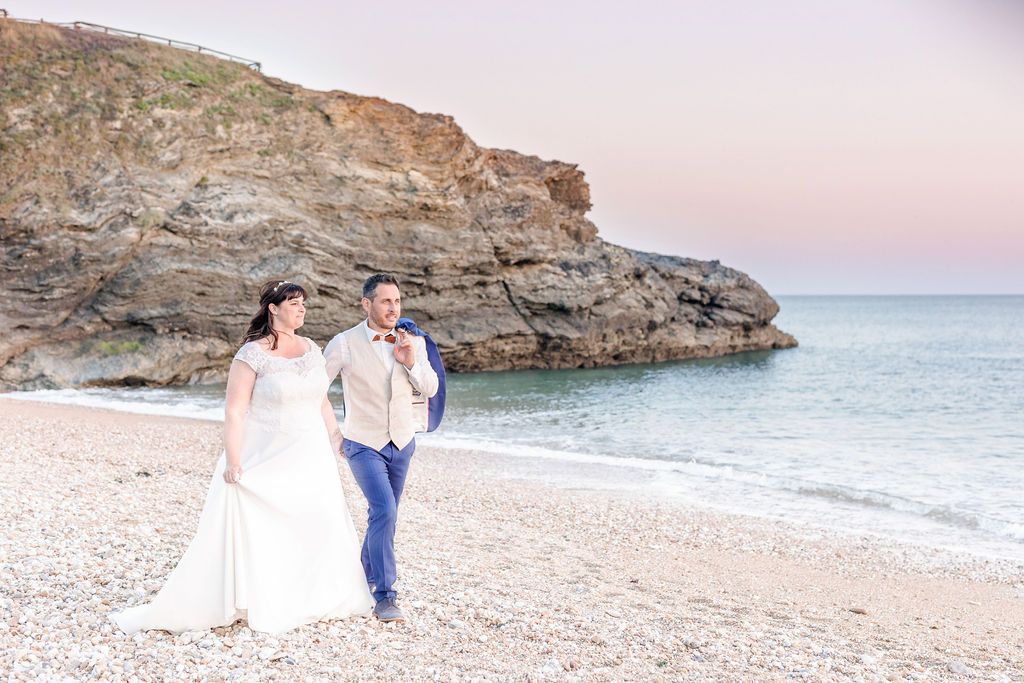 A beautiful beach wedding scene with the couple walking along the shoreline near rocky cliffs.
