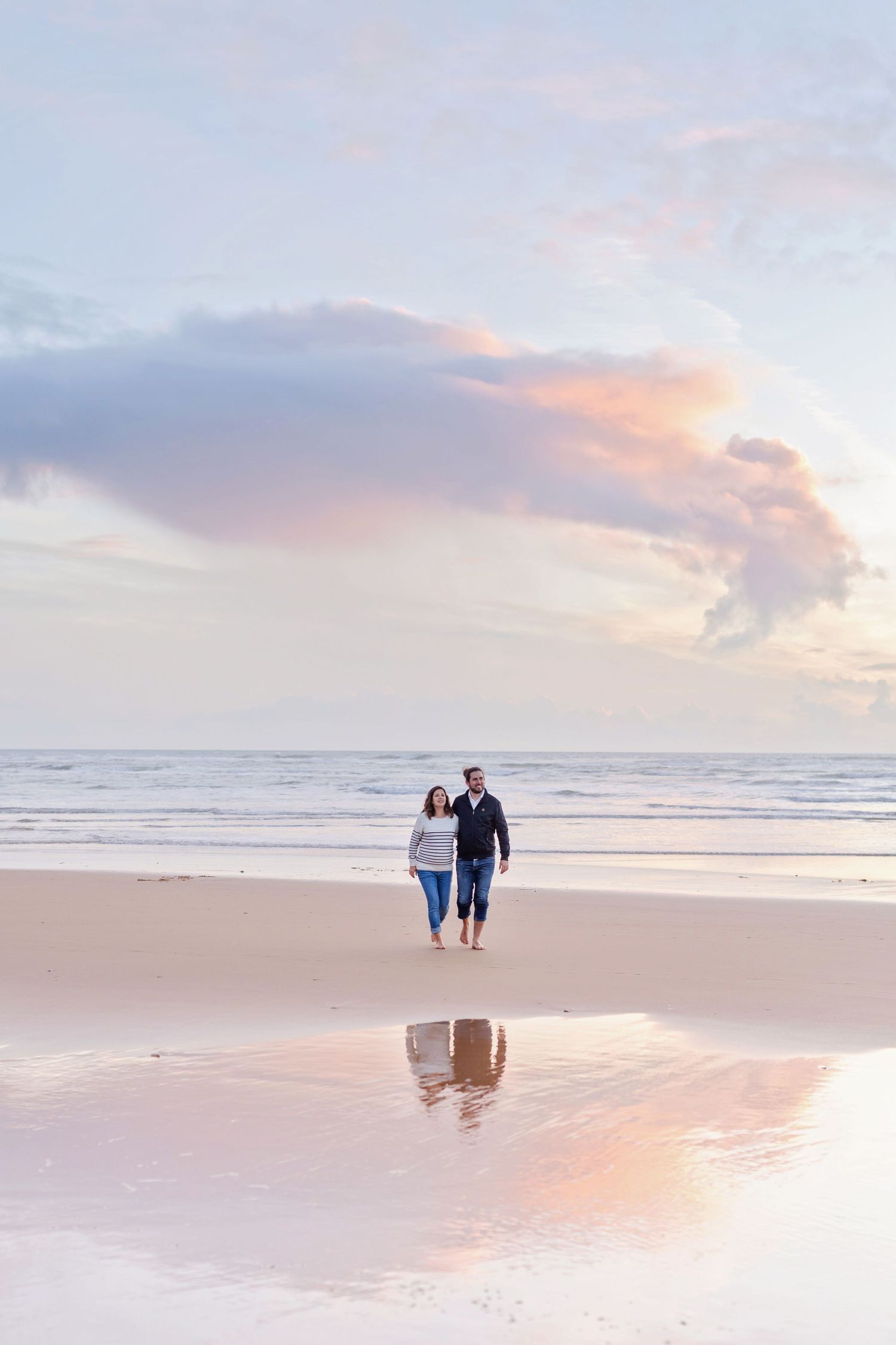A couple walks along a beach at sunset with their reflection in the wet sand.