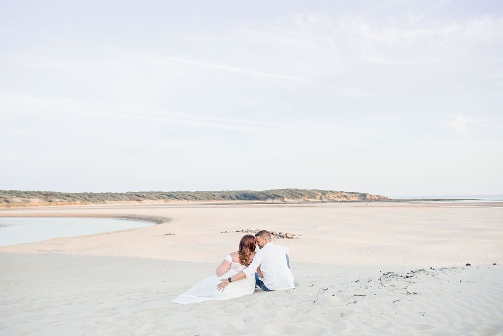 Romantic beach photoshoot with couple sitting on sandy shore at sunset.