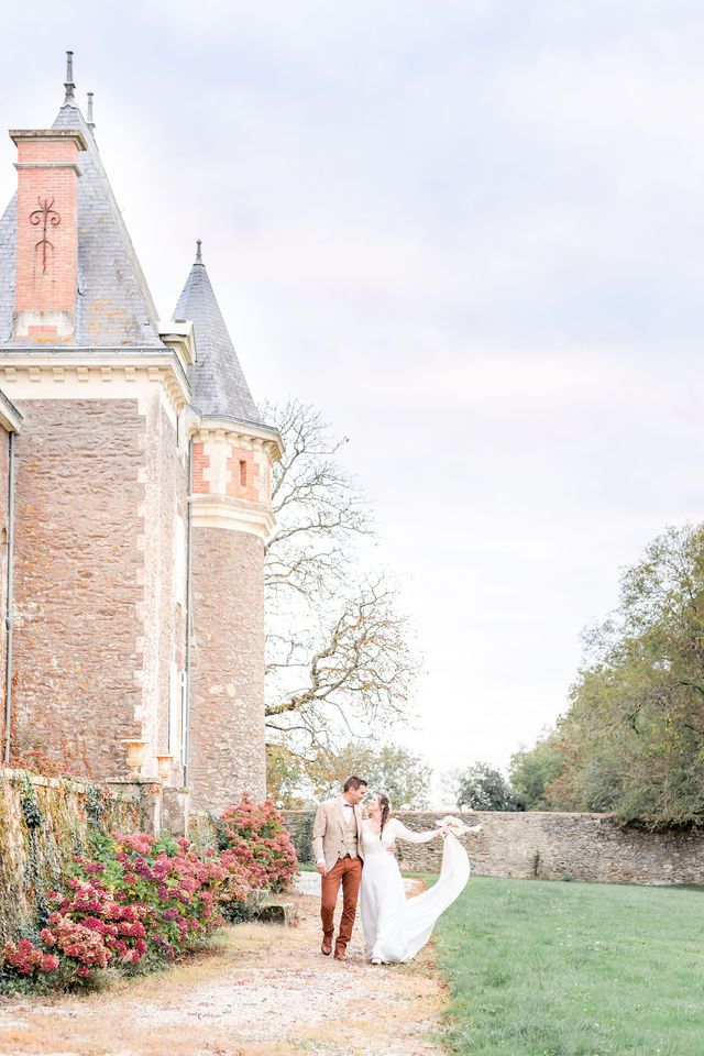Elegant wedding couple poses by stone castle walls with blooming red flowers.