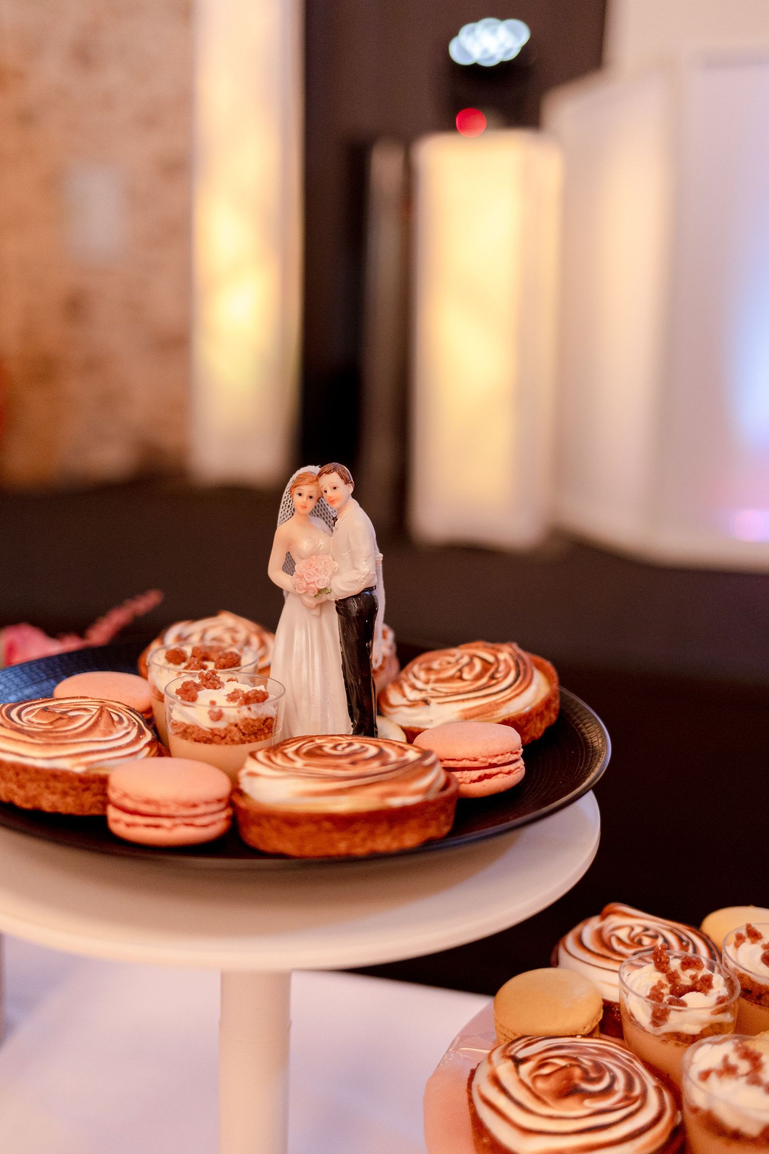 Wedding cake topper figurines displayed on a stand with decorated macarons and desserts.